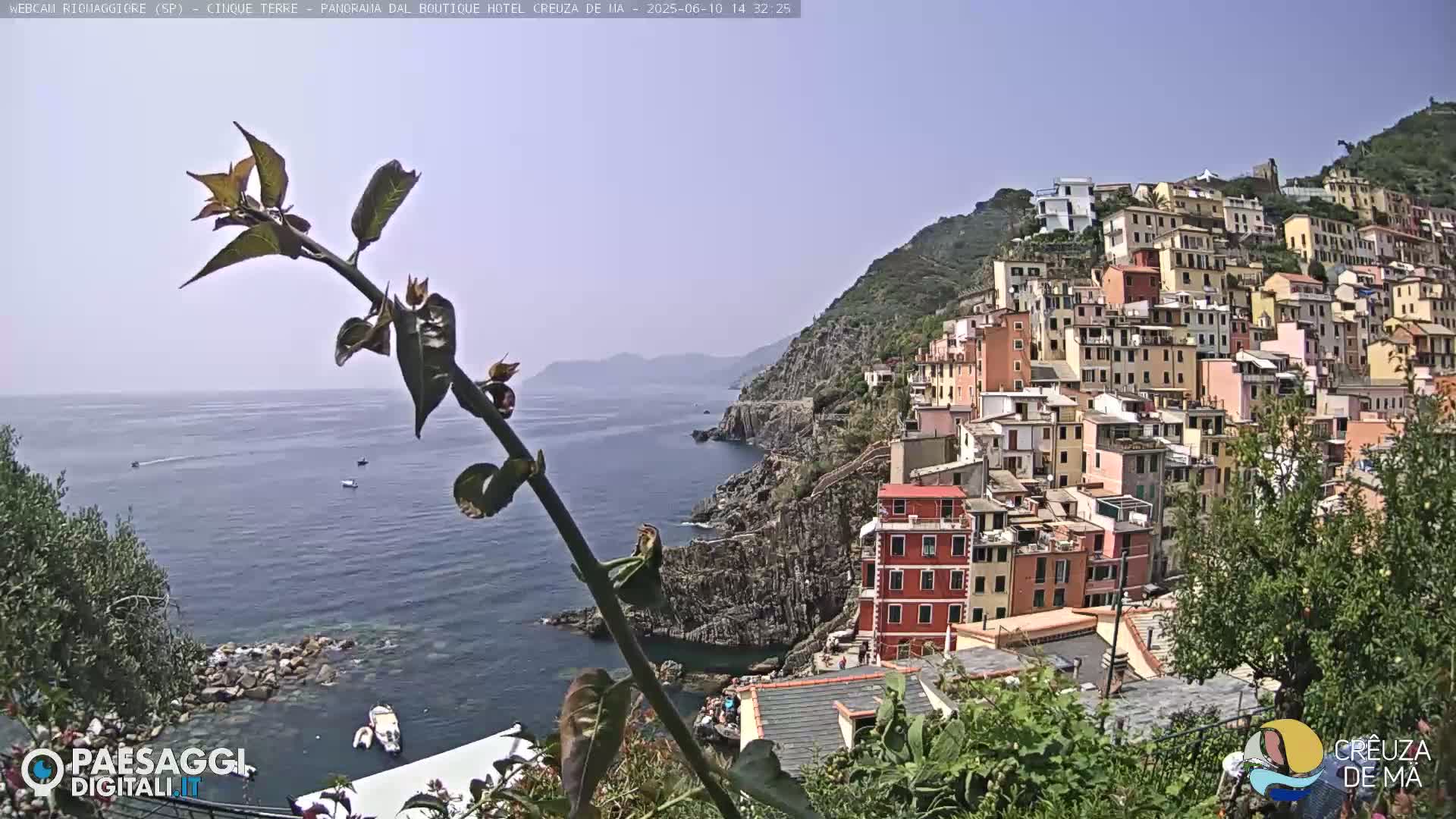 A coastal village of colorful buildings climbs a hillside overlooking a calm sea, with a few small boats visible on the water under a clear sky.
