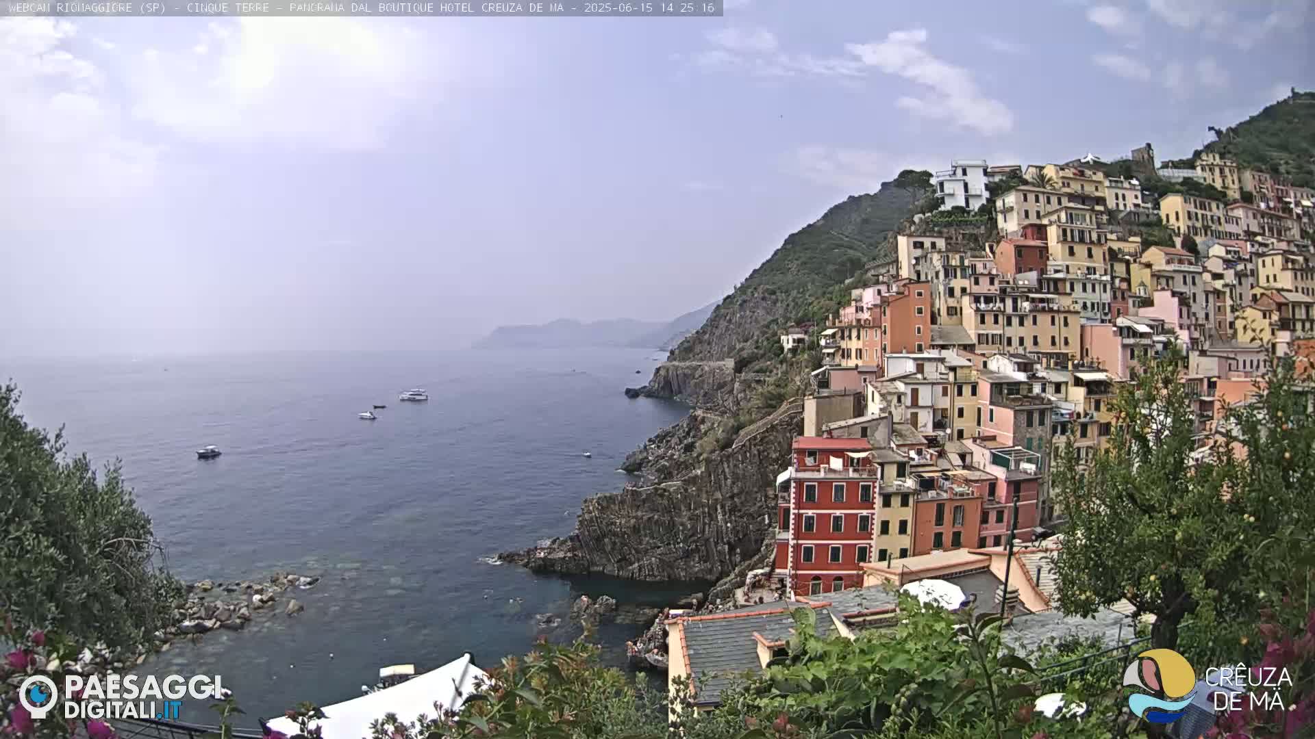 A coastal town with colorful buildings clinging to a steep hillside overlooks a calm sea with a few boats under a partly cloudy sky.