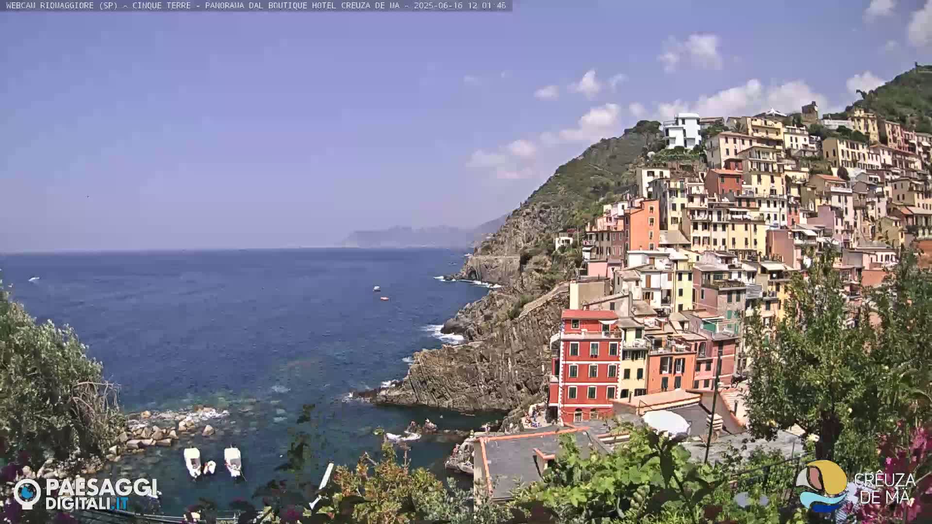 A colorful village clings to a steep cliffside overlooking a calm blue sea under a partly cloudy sky.