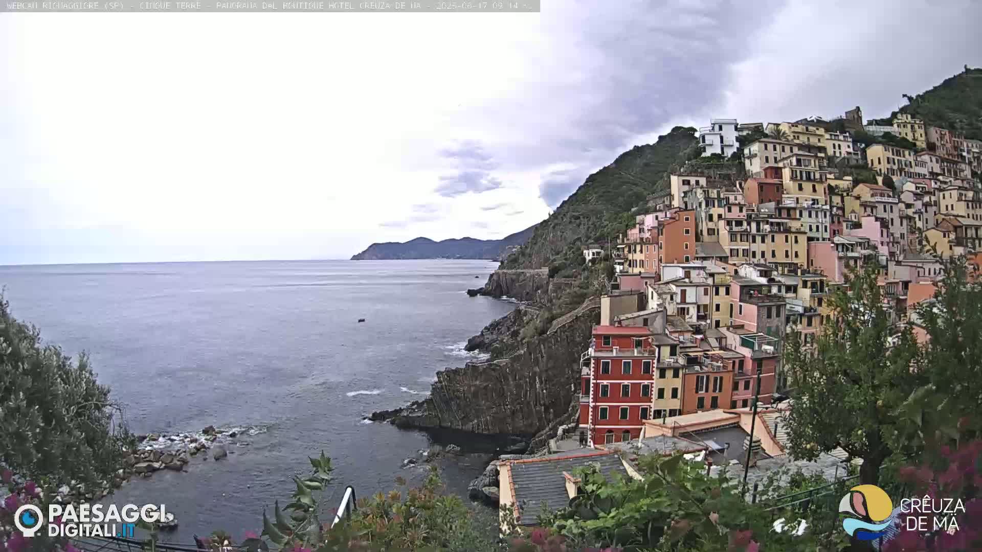 A coastal town with colorful buildings clinging to a steep hillside overlooks a calm sea under a cloudy sky.