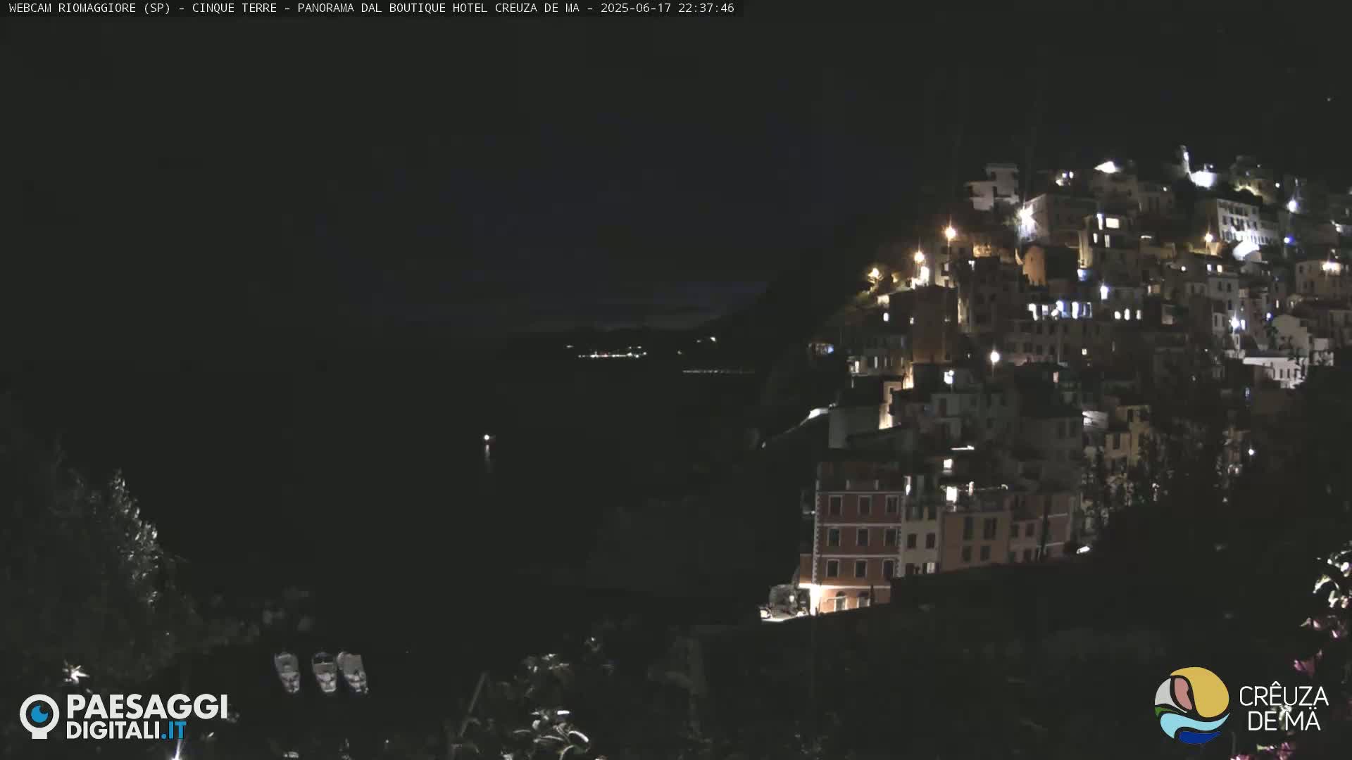 A nighttime view of a hillside town illuminated by lights, overlooking a dark body of water under a clear night sky.