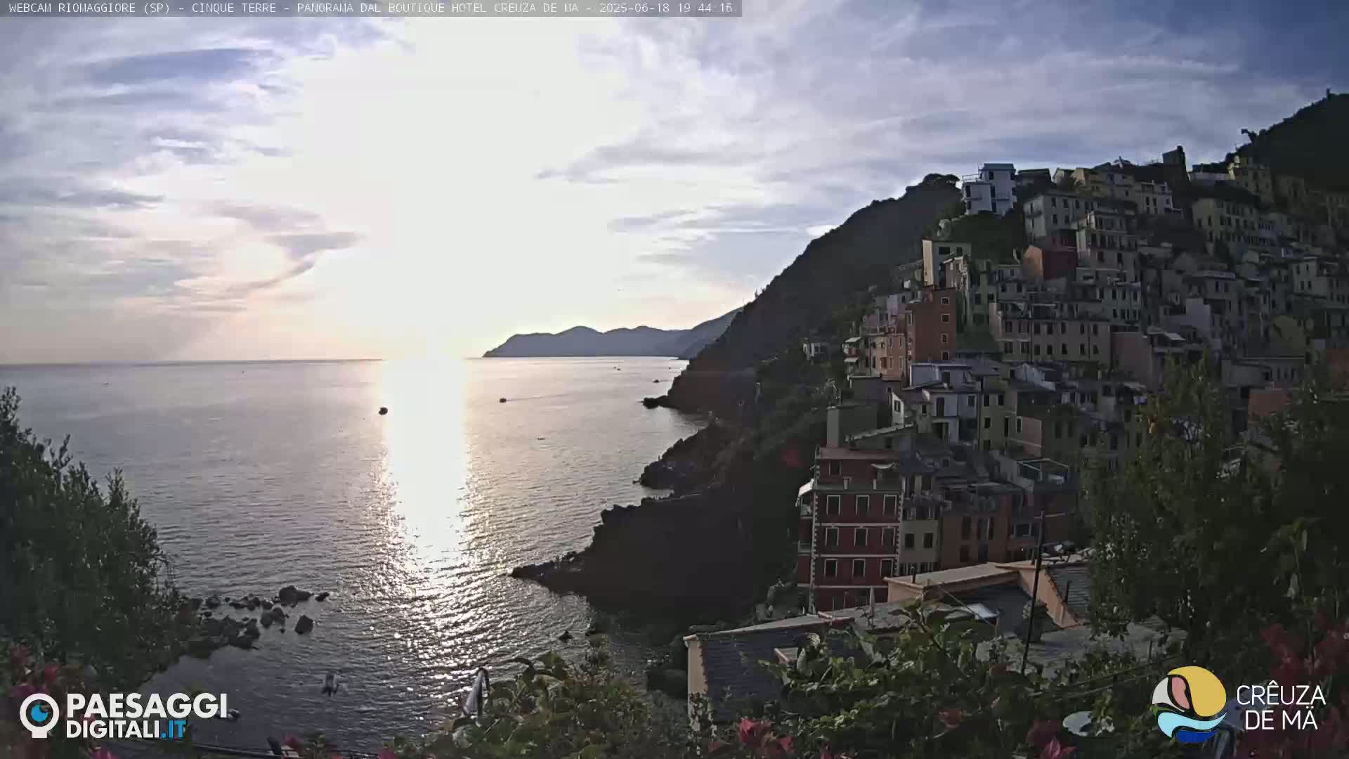 A seaside village clings to a cliffside overlooking a calm sea at sunset under a partly cloudy sky.