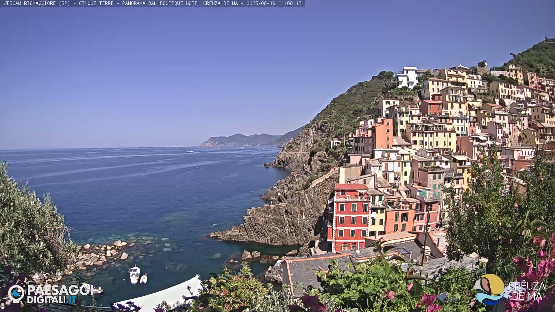 A colorful village clings to a hillside overlooking a calm blue sea under a clear sky.