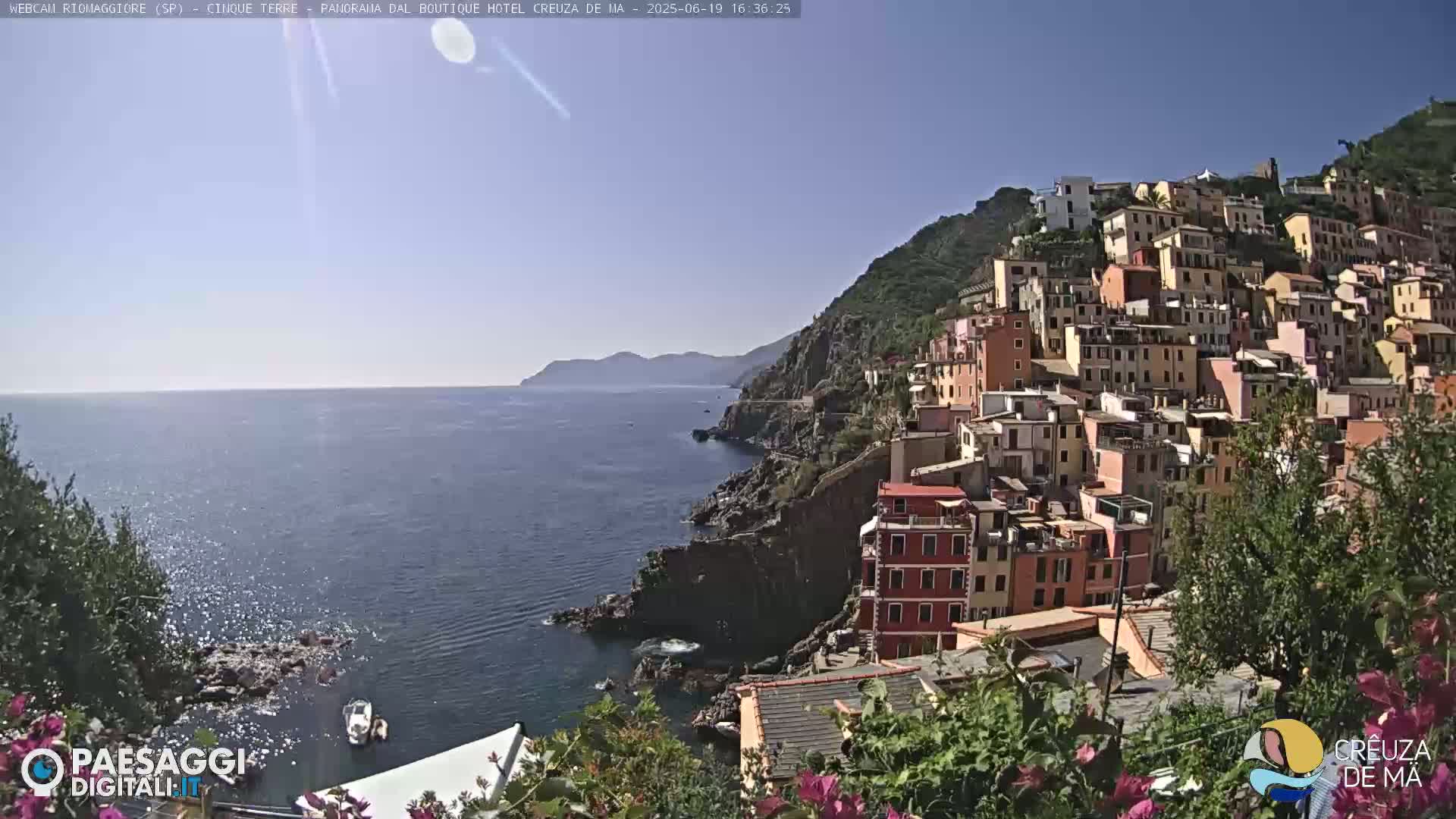 A colorful hillside village clings to a rocky coast overlooking a calm blue sea under a clear sunny sky.