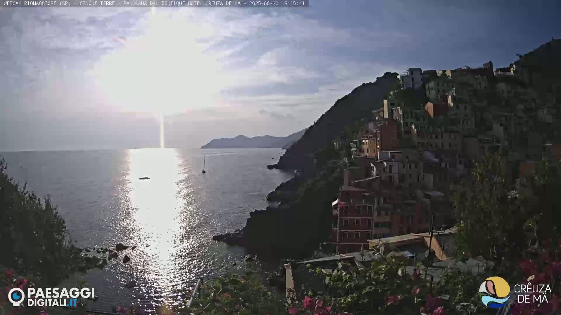 A seaside village clinging to a steep cliffside is bathed in the late afternoon sun, with the bright reflection of the sun shimmering on a calm sea.