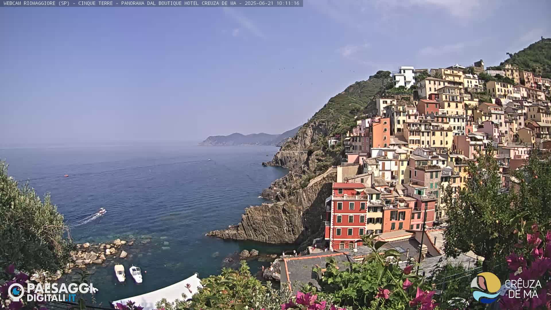 A colorful village clings to a steep, rocky hillside overlooking a calm blue sea, under a clear sunny sky.