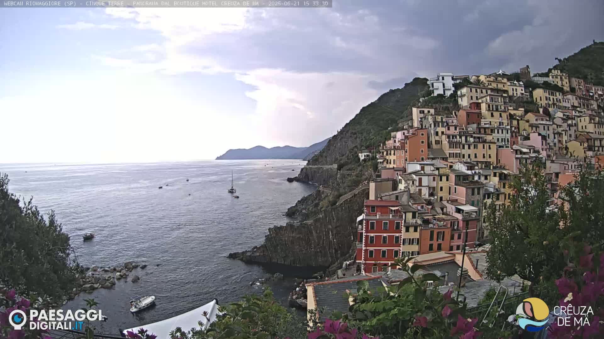A coastal village of colorful buildings clings to a steep hillside overlooking a calm sea under a partly cloudy sky.