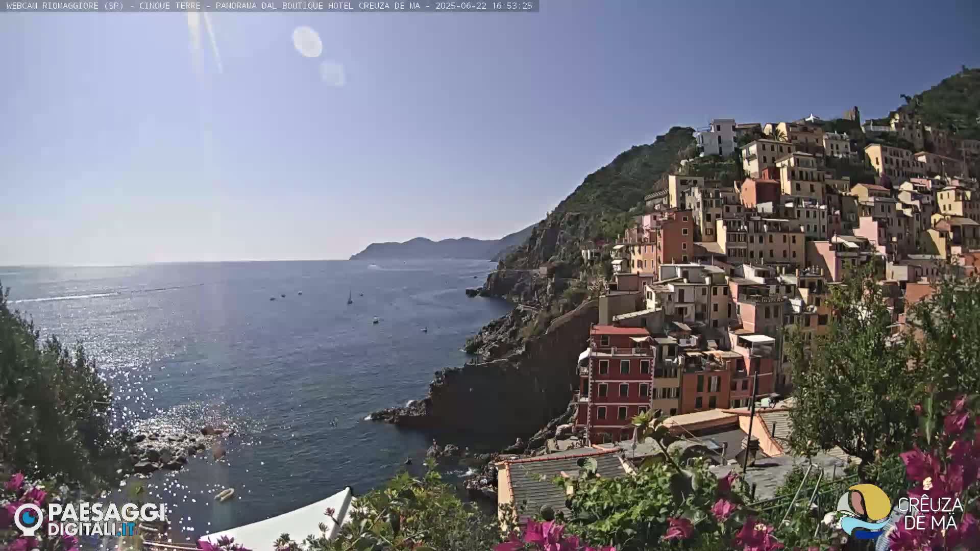 A colorful hillside village overlooks a calm, blue sea dotted with a few boats under a clear, sunny sky.