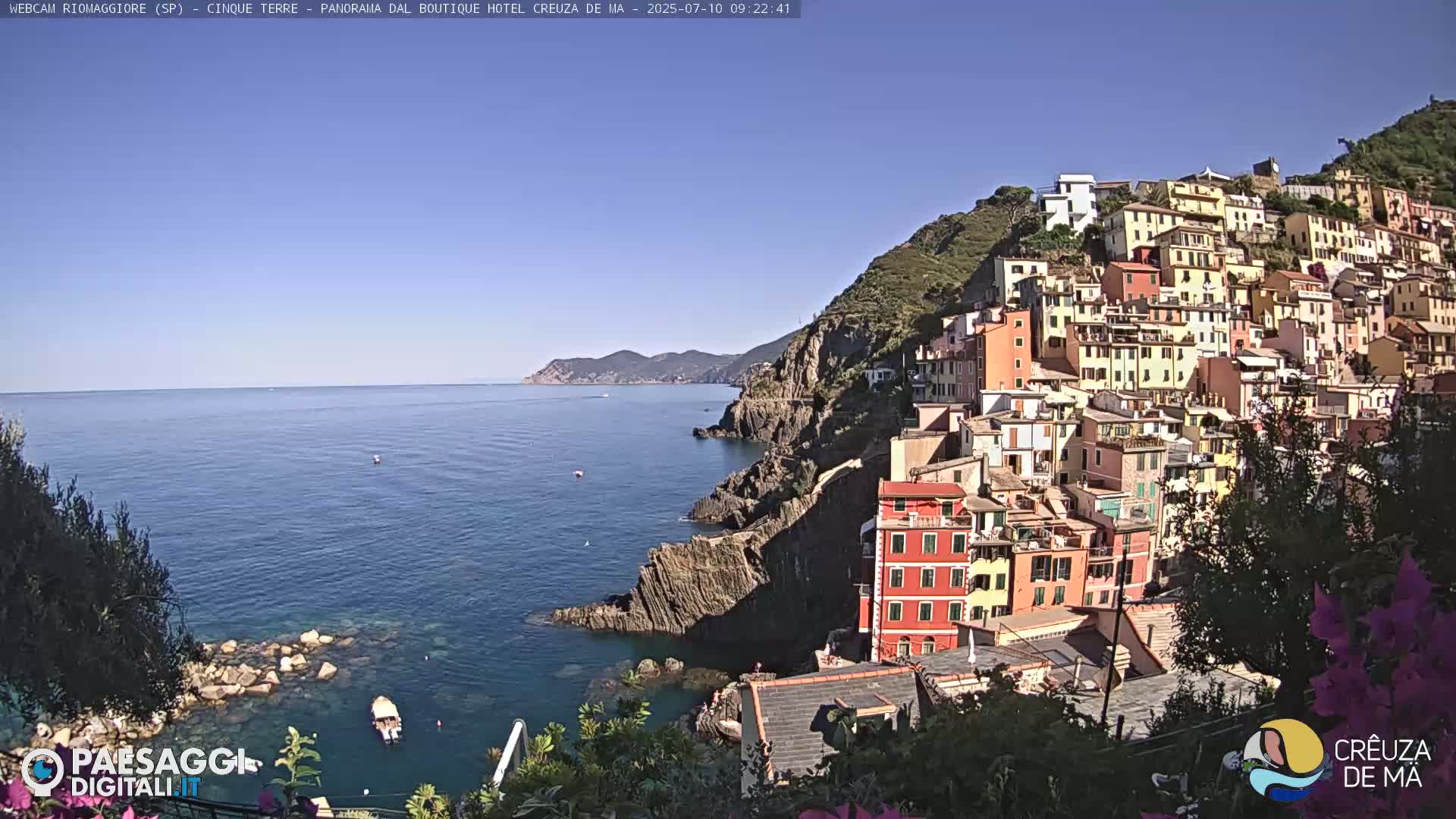 A colorful hillside village overlooks a calm, blue sea on a sunny day.