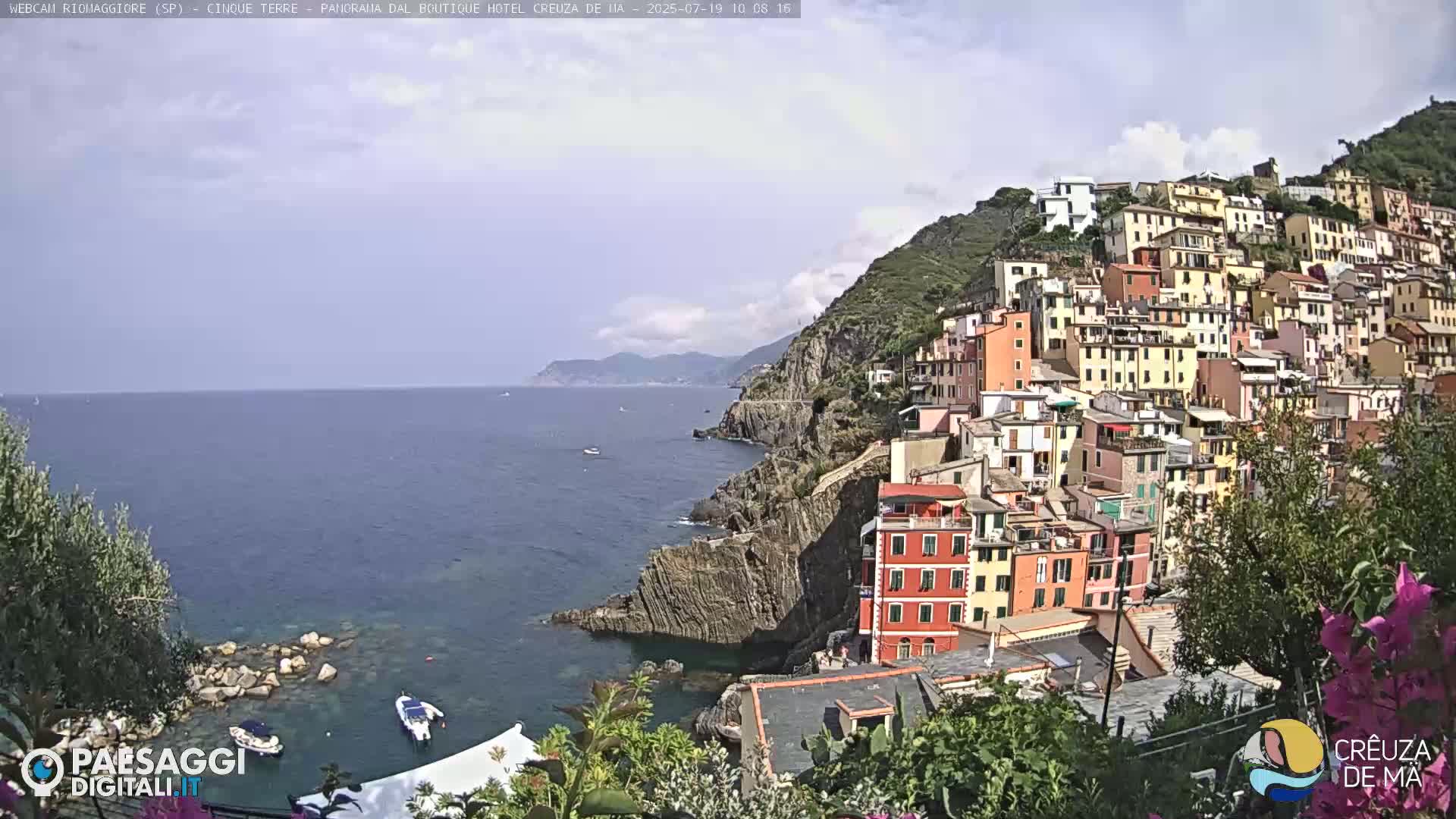 A colorful hillside village overlooks a calm blue sea on a partly cloudy day.