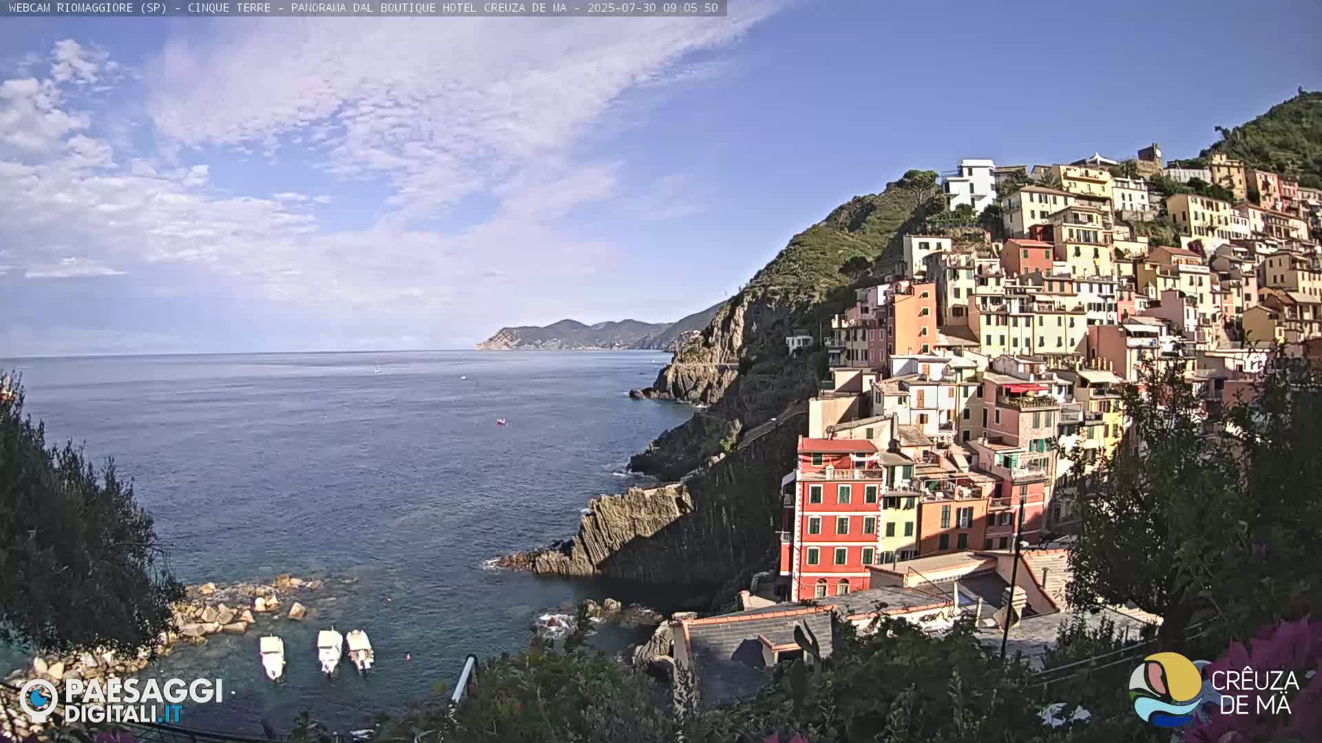 A colorful hillside village clings to a rocky coastline overlooking a calm, blue sea under a mostly sunny sky.