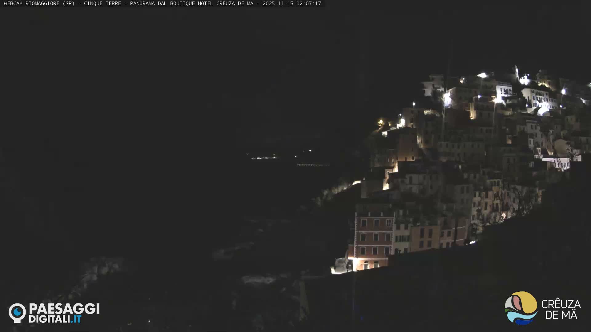 A brightly illuminated hillside village cascades down a slope toward a dark, calm sea with distant lights, all under a clear night sky.