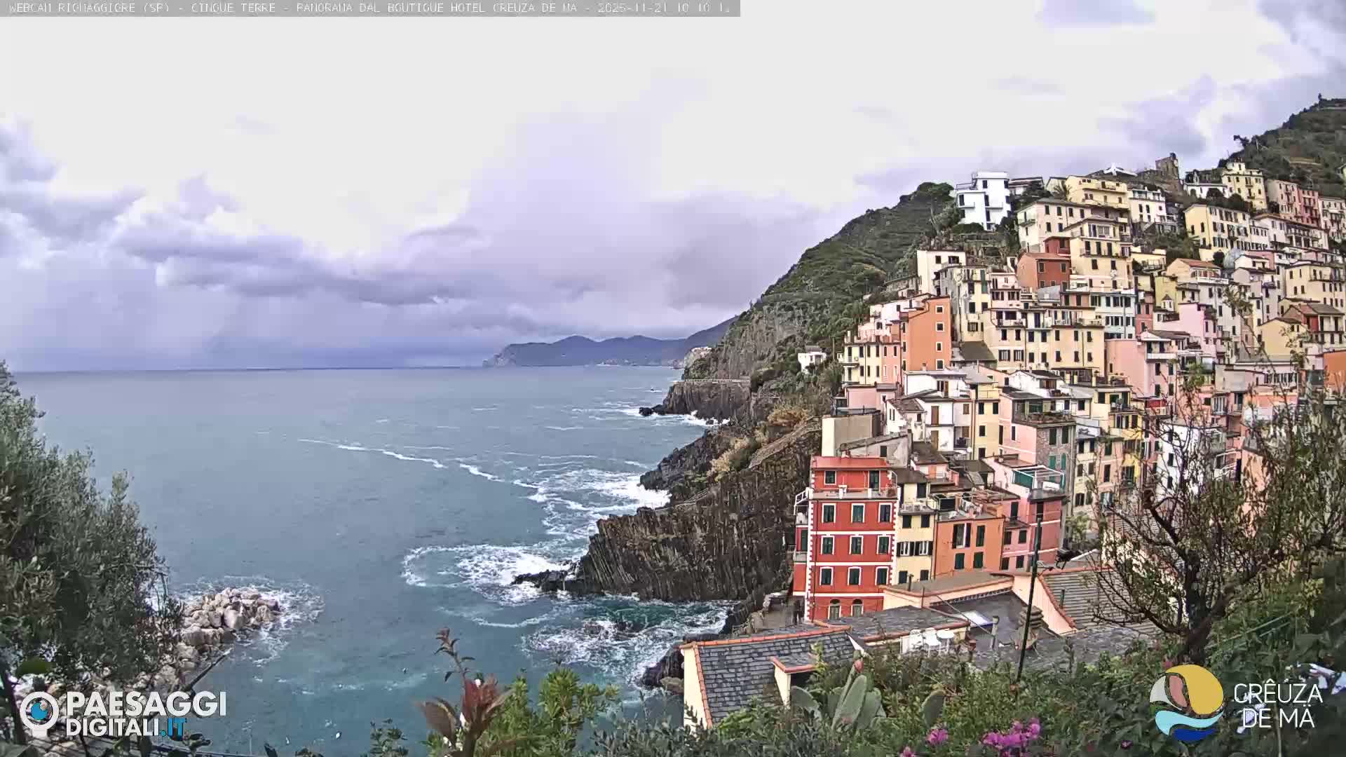 A colorful coastal town with densely packed buildings is nestled into a steep hillside overlooking a rocky shoreline with crashing waves under an overcast sky.