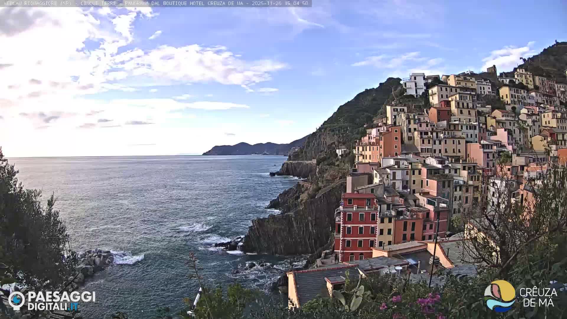 A colorful coastal town with densely packed buildings is nestled into a steep hillside overlooking a rocky shoreline with crashing waves under an overcast sky.