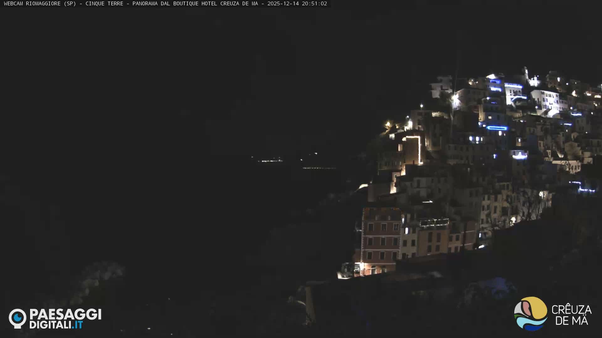 The image shows a vibrant, colorful town built into a steep, green hillside overlooking a rocky coastline and the sea under an overcast sky.