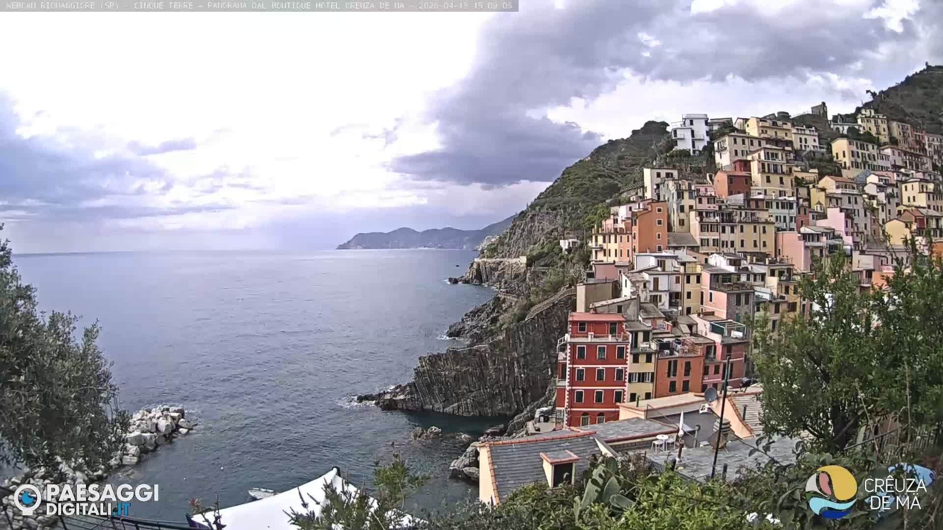 The image shows a vibrant, colorful town built into a steep, green hillside overlooking a rocky coastline and the sea under an overcast sky.