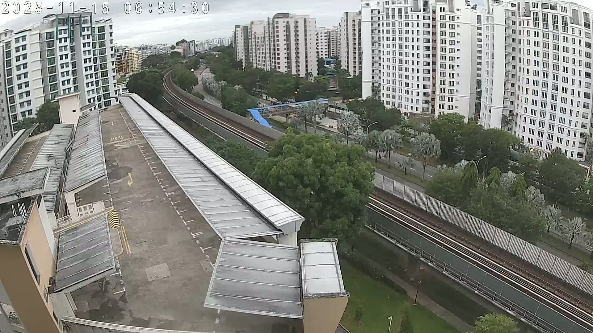 An elevated view of a modern city shows a long, covered walkway and parking area in the foreground, with elevated train tracks curving through green spaces, surrounded by numerous tall residential buildings, all under a heavily overcast sky.