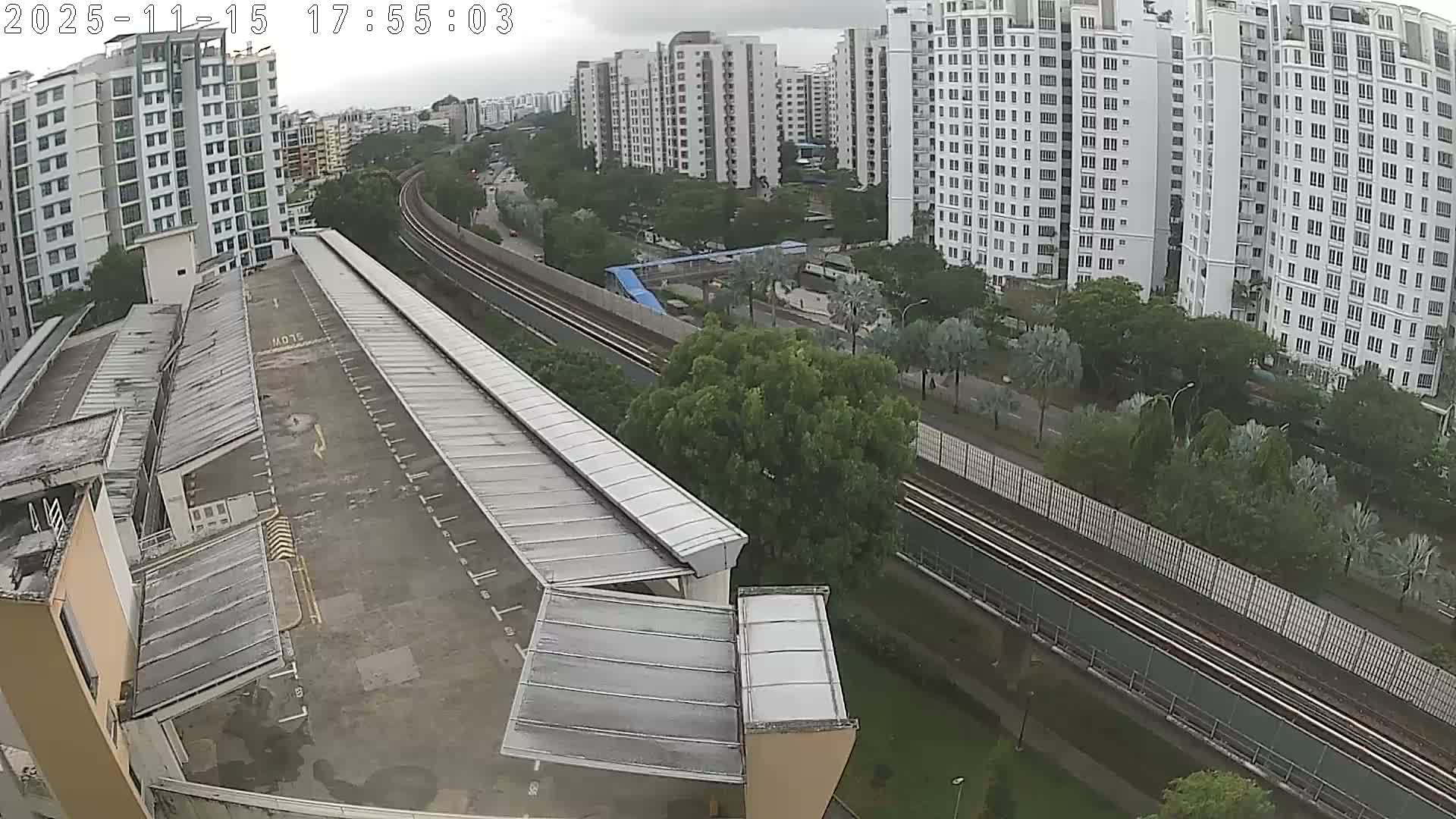 An elevated train track cuts through a dense urban environment dominated by numerous high-rise residential buildings and abundant tropical foliage, all under a heavily overcast sky.