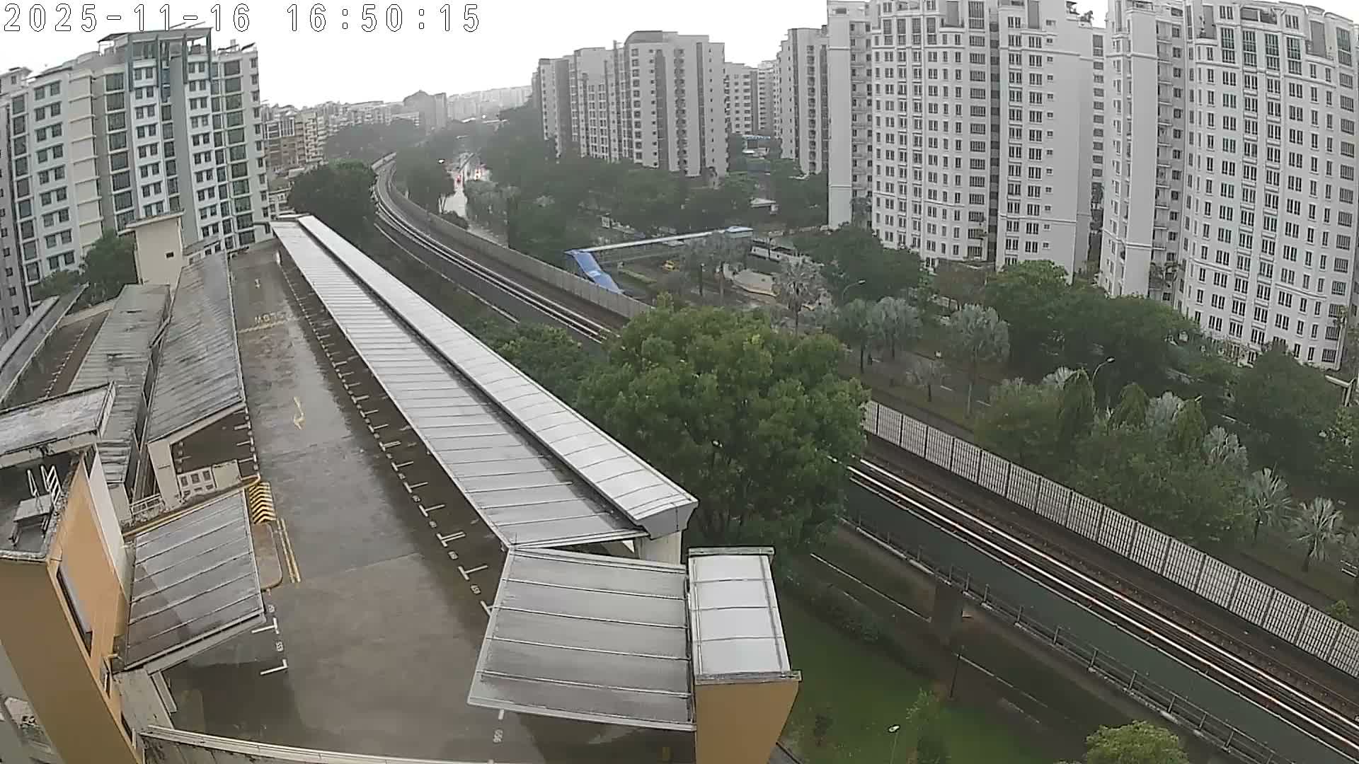 An elevated view showcases a modern cityscape featuring numerous white high-rise residential buildings, a rain-soaked multi-level car park with an extensive covered platform, and elevated train tracks weaving through abundant green trees under a heavily overcast and rainy sky.