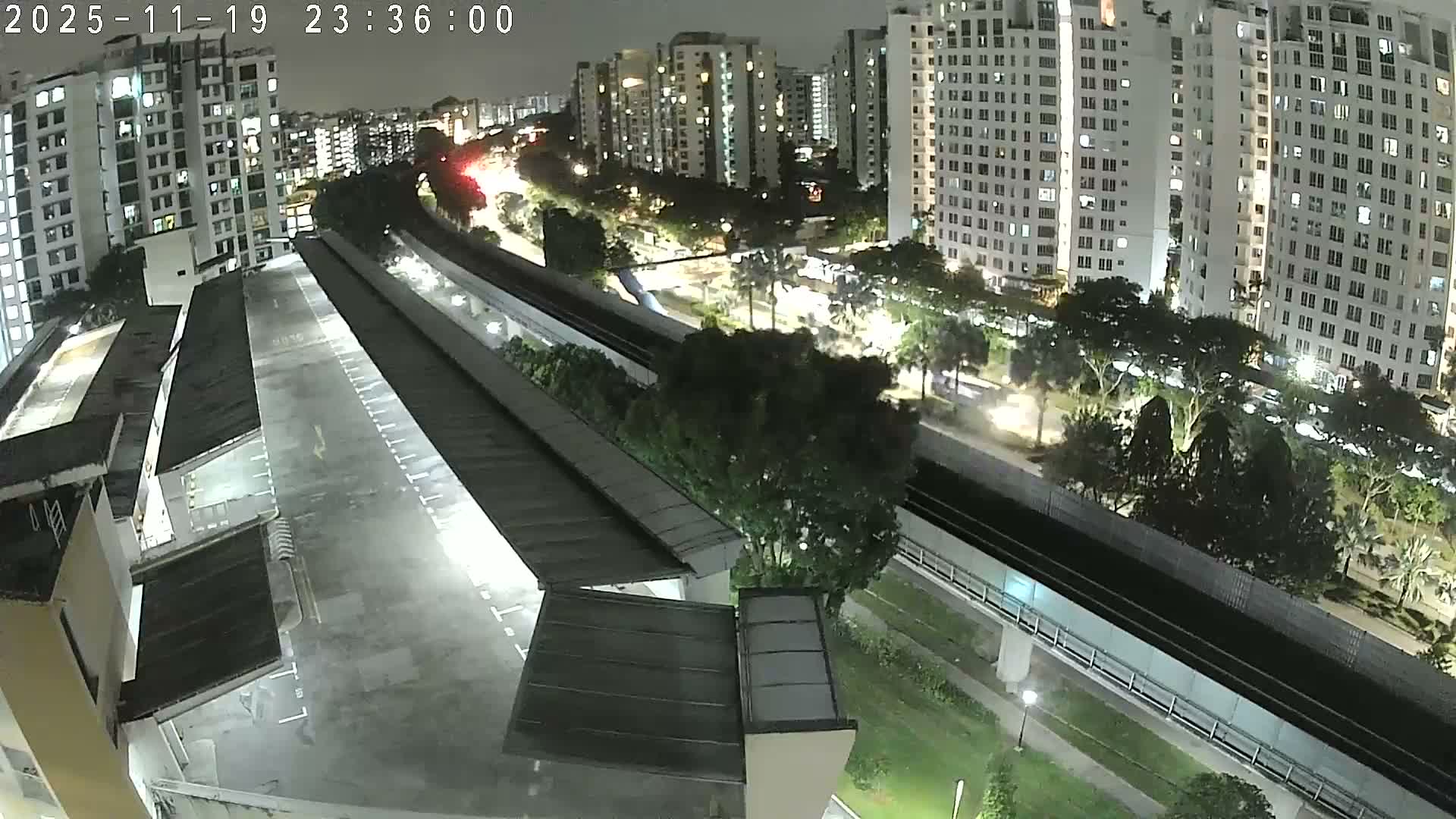 An elevated view showcases a modern cityscape featuring numerous white high-rise residential buildings, a rain-soaked multi-level car park with an extensive covered platform, and elevated train tracks weaving through abundant green trees under a heavily overcast and rainy sky.