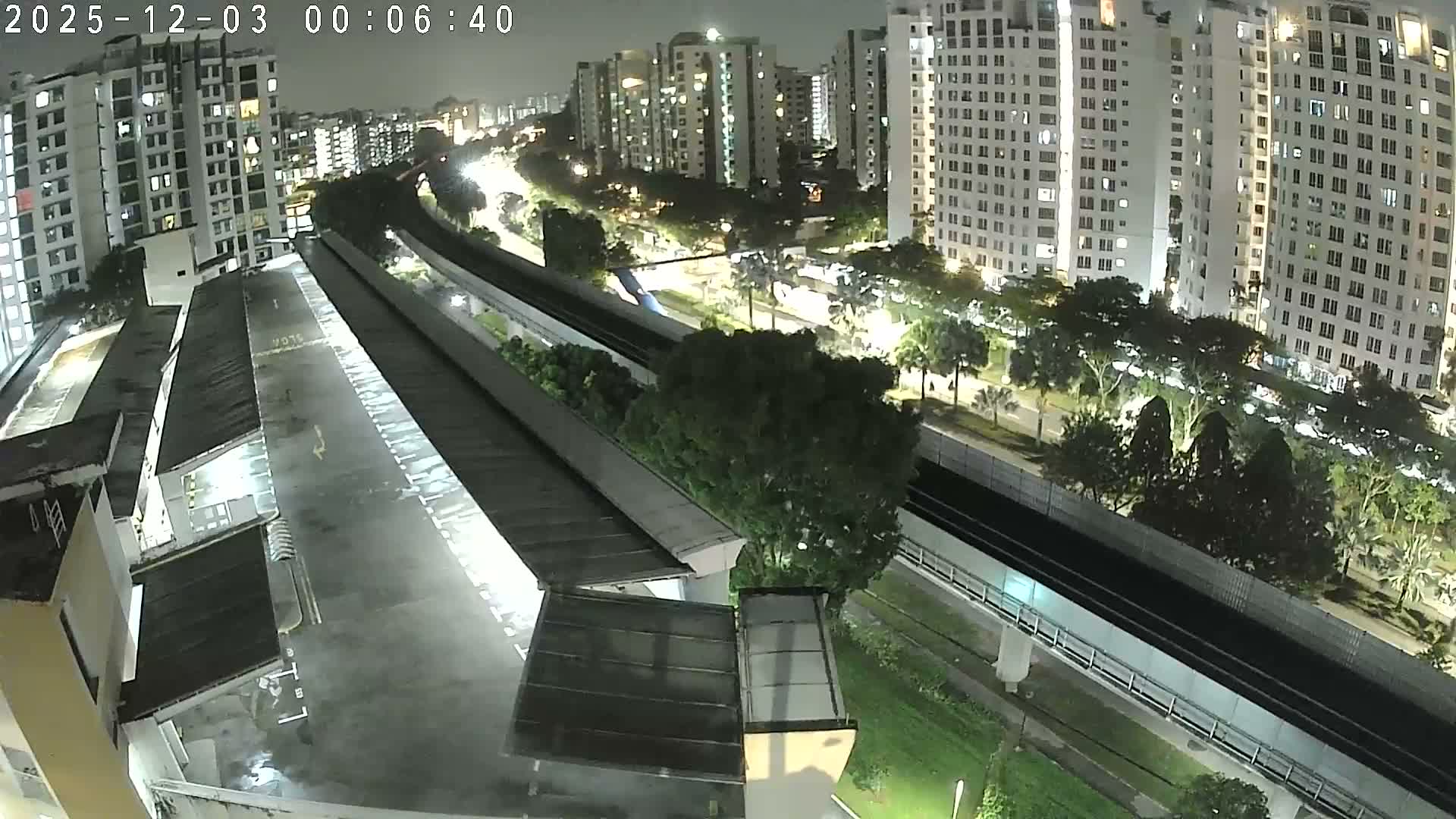 An elevated view at night captures a dense urban environment featuring a damp, reflective foreground structure, an elevated train track, and numerous brightly lit high-rise buildings overlooking streets with sparse traffic and abundant trees, all under overcast and damp conditions.