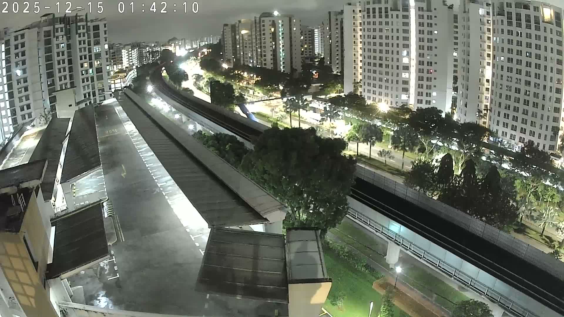 An elevated view captures a city scene with numerous tall residential buildings, an elevated train line snaking through lush greenery, and a covered parking structure in the foreground, all bathed in the warm, hazy light of the late afternoon.