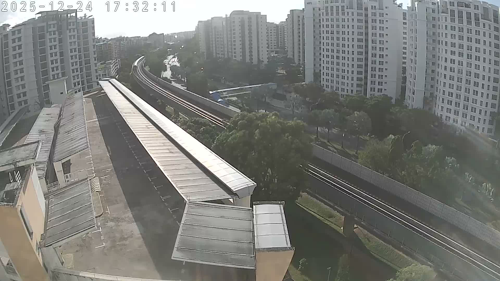 An elevated view captures a city scene with numerous tall residential buildings, an elevated train line snaking through lush greenery, and a covered parking structure in the foreground, all bathed in the warm, hazy light of the late afternoon.