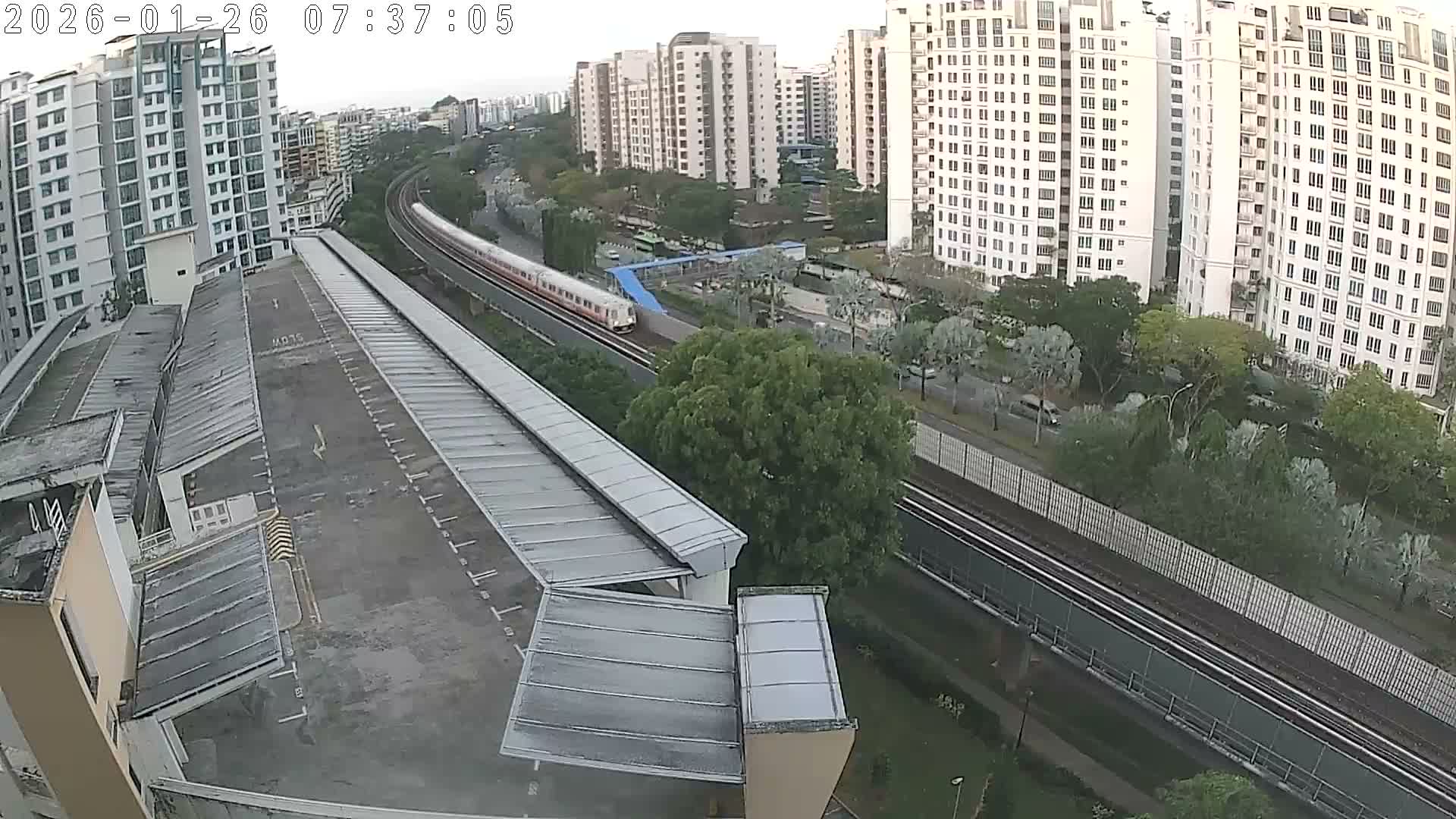 An elevated view on an overcast day captures a white and red train traversing curved tracks through a bustling urban landscape filled with numerous high-rise buildings, lush greenery, and a large multi-level covered structure in the foreground.