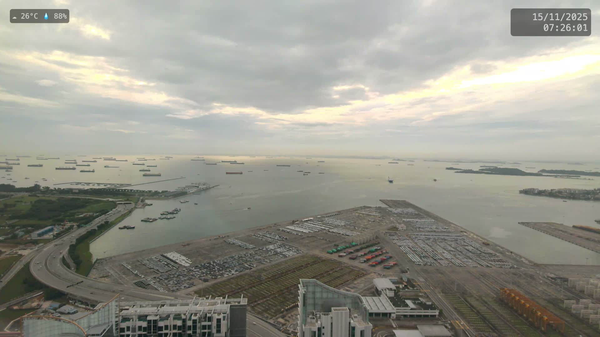 Under an overcast sky, an aerial view captures a vast strait dotted with numerous cargo ships, bordered by a coastal highway and a sprawling port facility with large lots of parked vehicles.