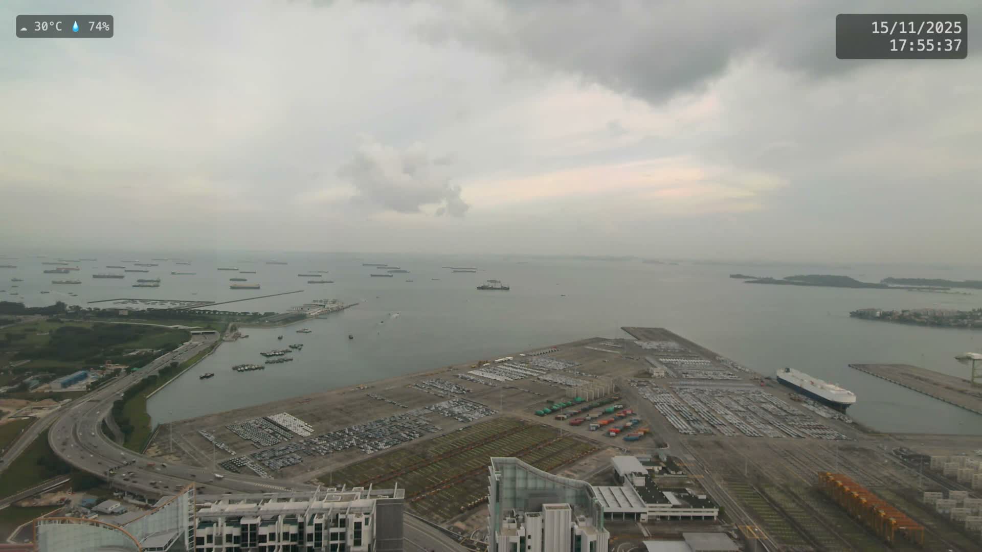 An expansive aerial view reveals a busy port with numerous ships anchored in the bay, extensive car storage lots, and a coastal highway, all beneath an overcast sky.