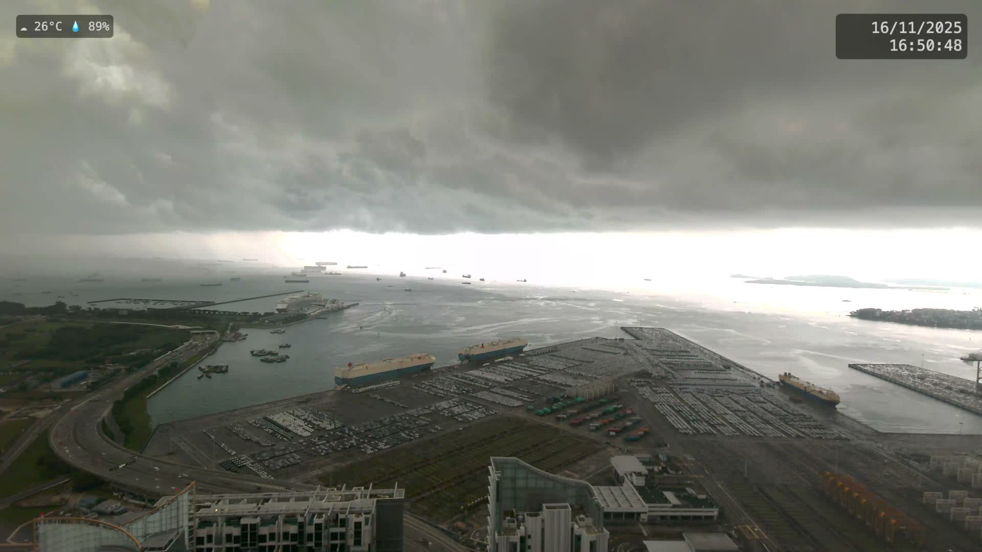 An expansive port facility, bustling with ships and filled with rows of new cars, is seen under a dark, stormy, and heavily overcast sky.