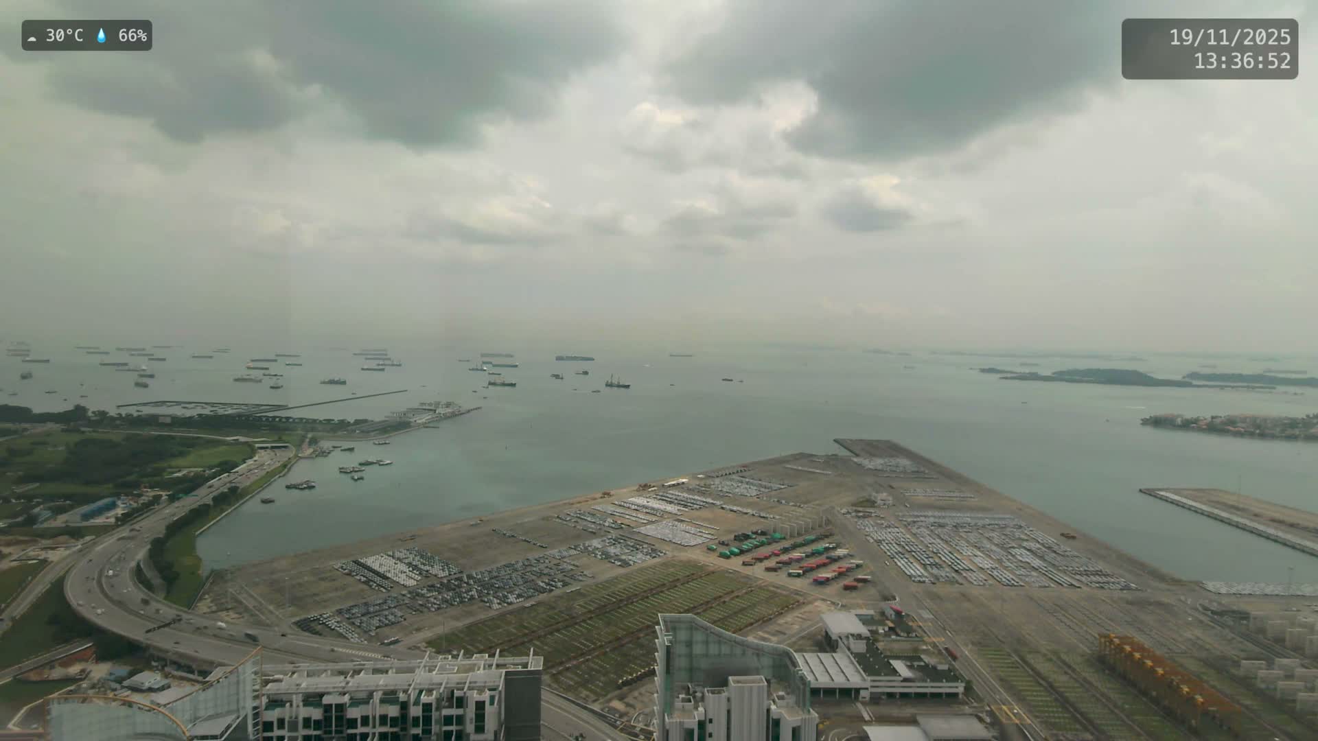 An expansive port facility, bustling with ships and filled with rows of new cars, is seen under a dark, stormy, and heavily overcast sky.