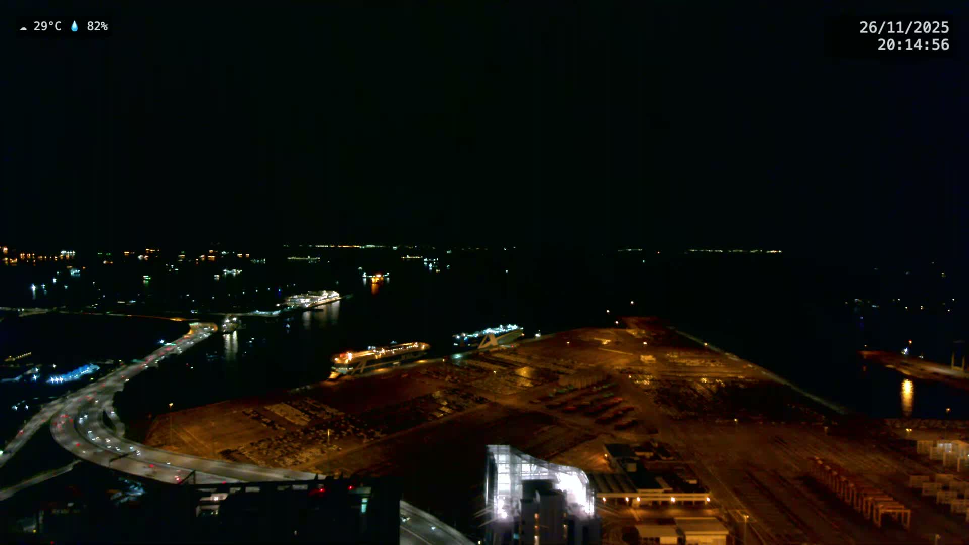 On an overcast day, an aerial view reveals a sprawling port with thousands of new cars parked in vast lots, a multi-lane coastal highway, and countless cargo ships anchored across a wide bay with distant islands.