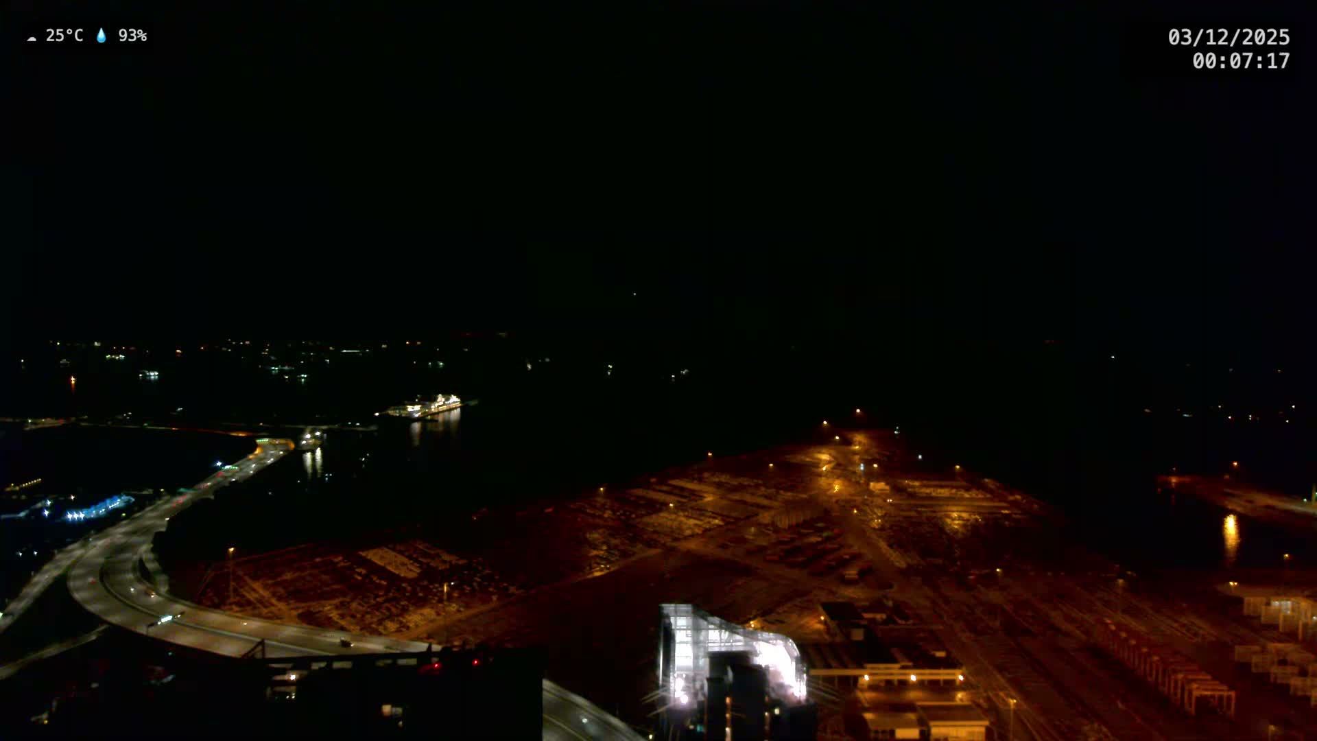 An aerial view at night captures a vast industrial port and harbor area, featuring a brightly lit cruise ship, a curving illuminated highway, and numerous facility lights, all under a clear, warm, and humid sky.