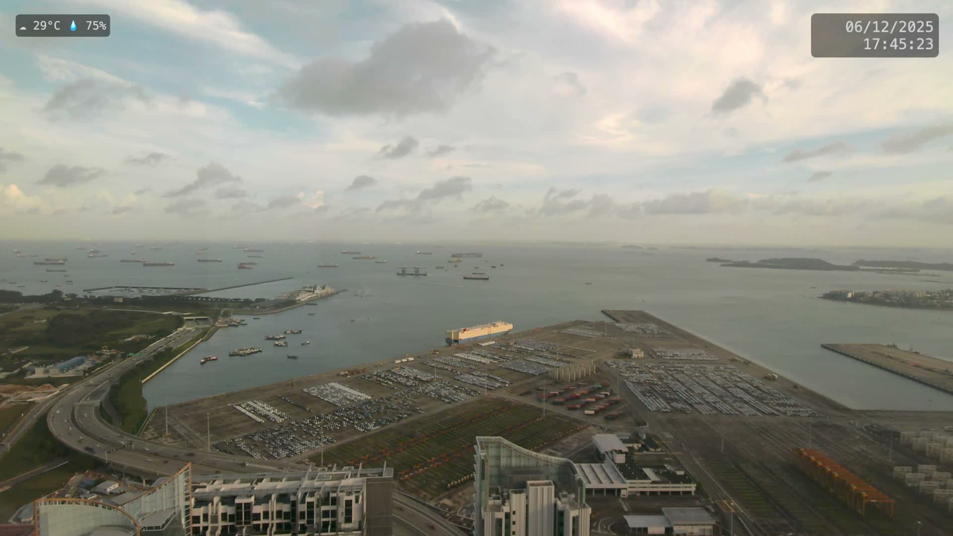 A wide aerial view under overcast and hazy skies reveals a vast body of water dotted with numerous cargo ships, a sprawling port facility featuring extensive car storage lots and a docked car carrier, alongside winding coastal highways and urban structures.