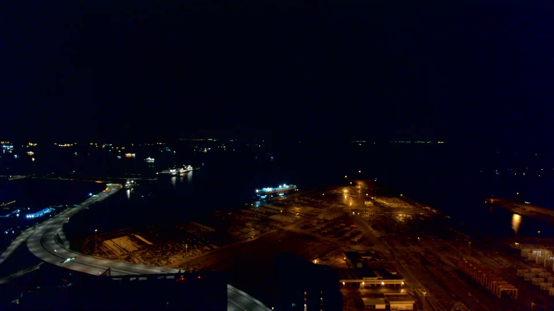 A wide aerial view under overcast and hazy skies reveals a vast body of water dotted with numerous cargo ships, a sprawling port facility featuring extensive car storage lots and a docked car carrier, alongside winding coastal highways and urban structures.