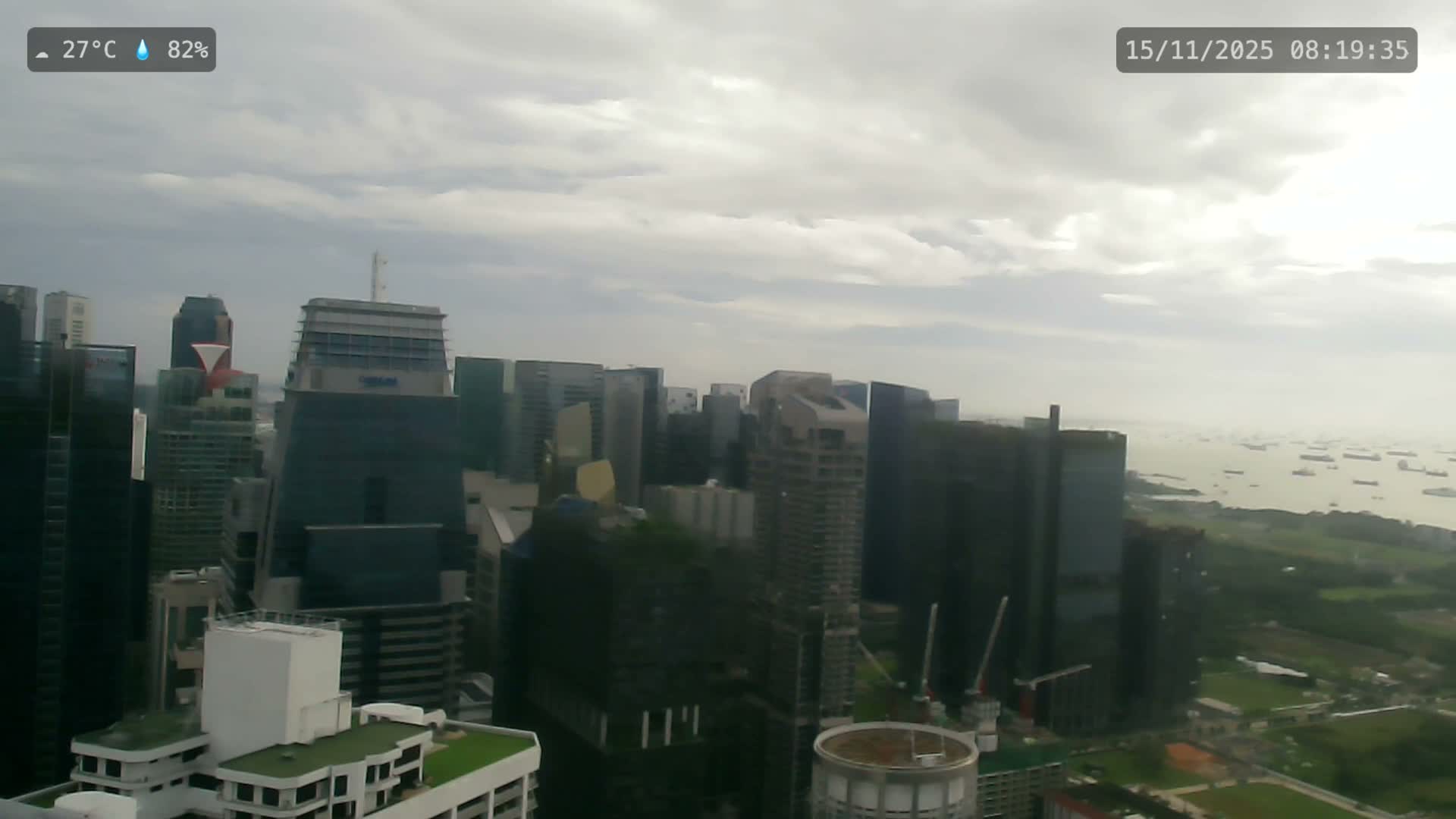 A panoramic view showcases a sprawling cityscape of numerous skyscrapers and ongoing construction leading to a busy bay filled with ships, all under a heavily overcast and cloudy sky.