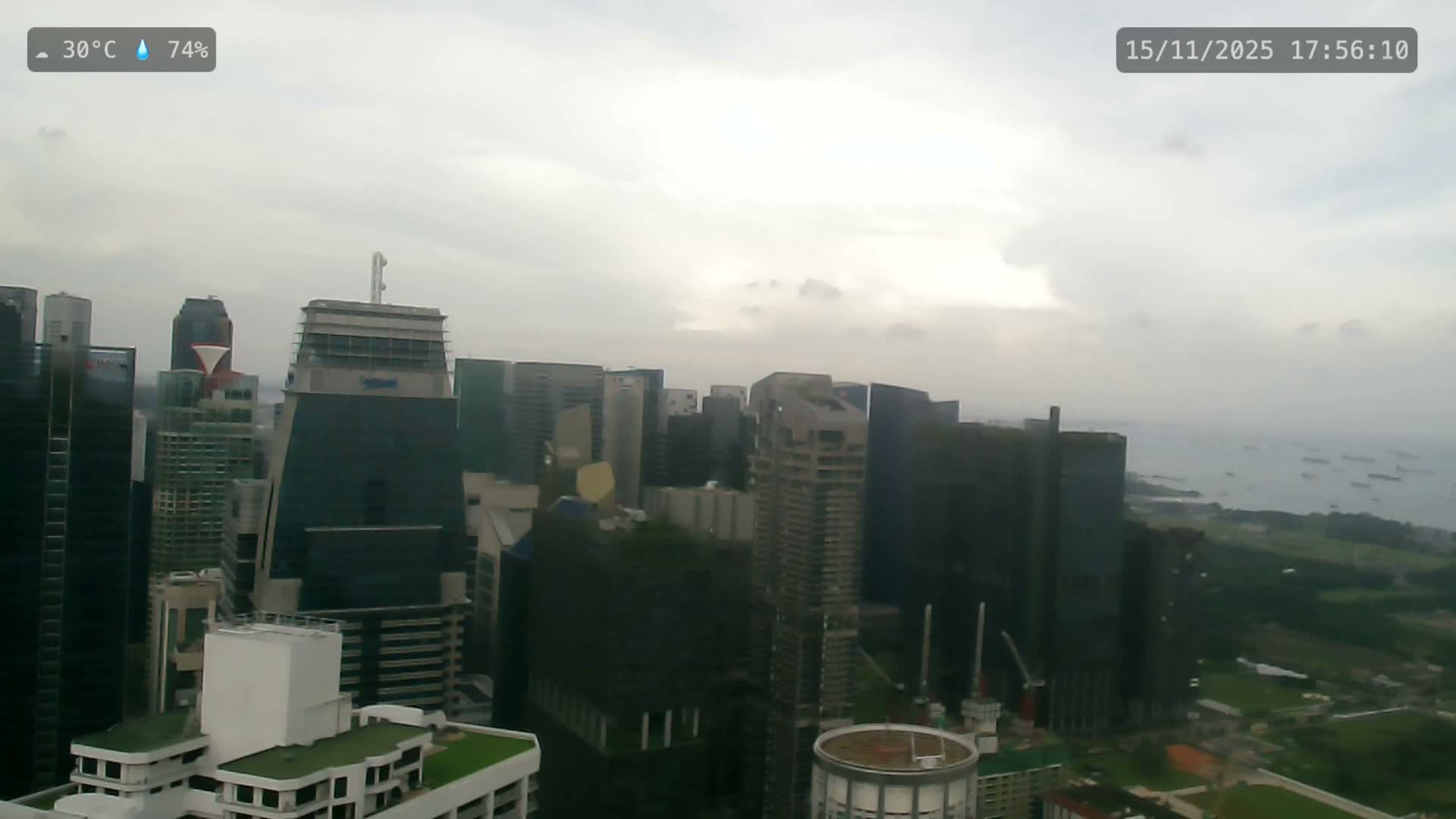 An aerial view reveals a dense cityscape of high-rise buildings extending towards a hazy sea dotted with numerous ships, all under a bright, overcast sky.