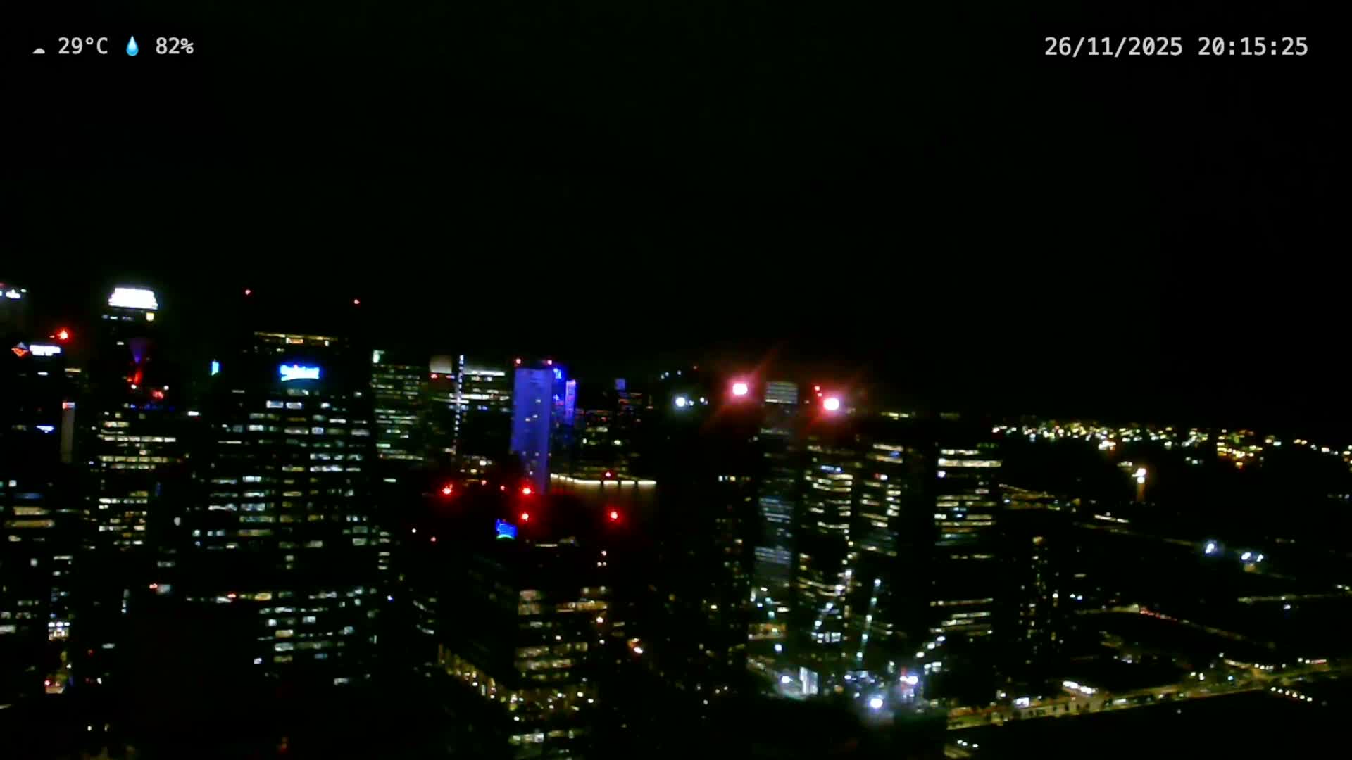 An expansive view of a modern city under an overcast sky, featuring numerous high-rise buildings with ongoing construction, and a vast body of water dotted with many ships in the hazy distance.