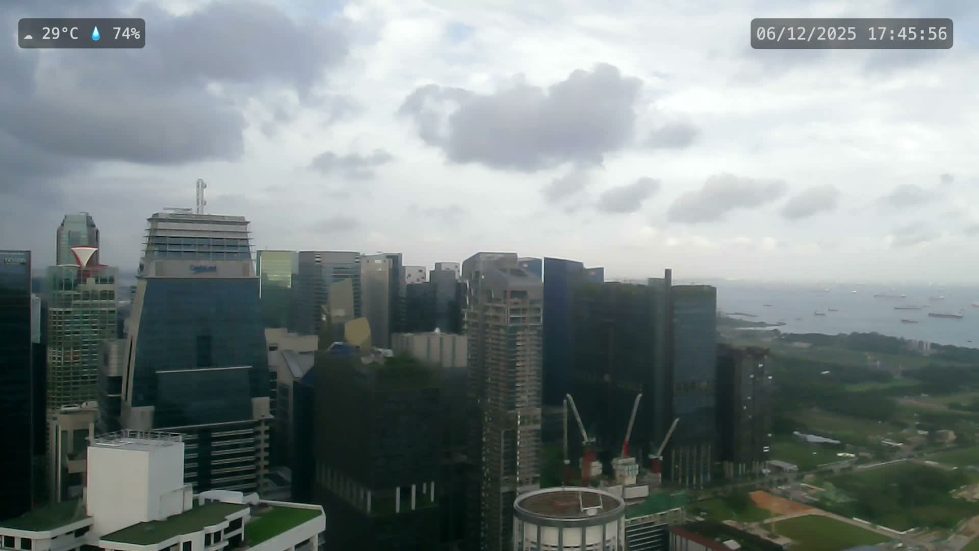 Under an overcast and hazy sky, a dense modern cityscape features numerous tall buildings and construction cranes, with a wide body of water dotted by many ships visible in the distant background.