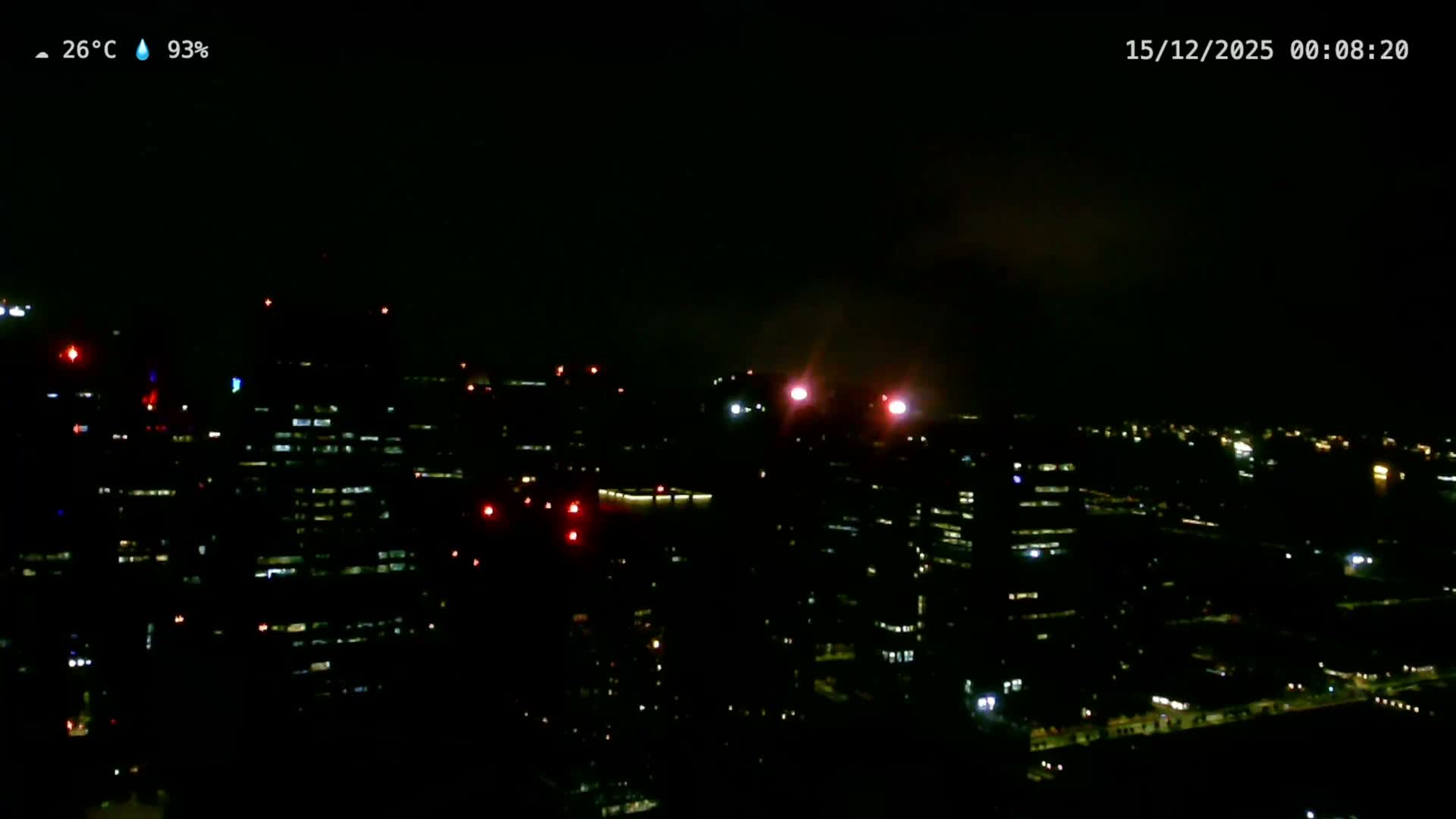 Under an overcast and hazy sky, a dense modern cityscape features numerous tall buildings and construction cranes, with a wide body of water dotted by many ships visible in the distant background.