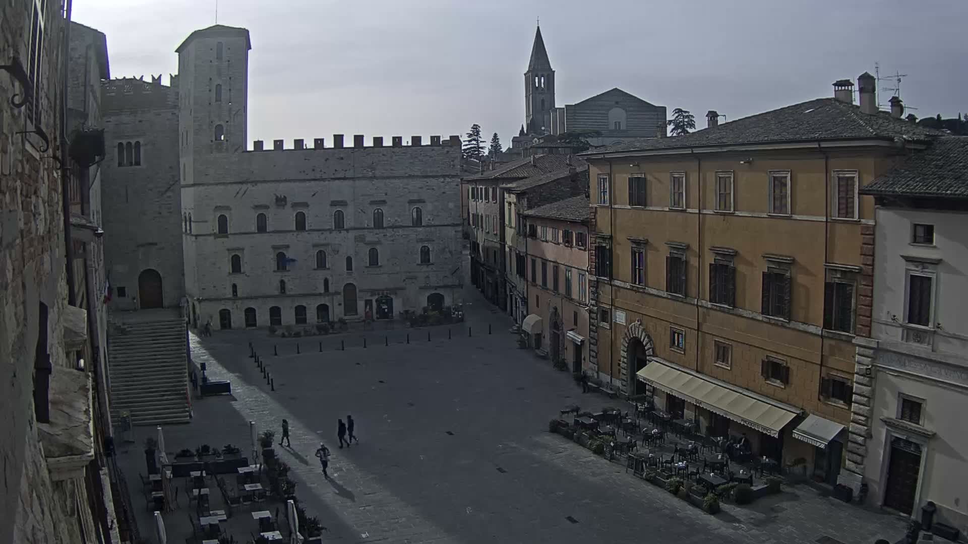 An overcast daytime view reveals a historic European town square with ancient stone buildings, a prominent tower on the left, a distant church spire, and a few people walking across the plaza while an outdoor cafe operates on the right.