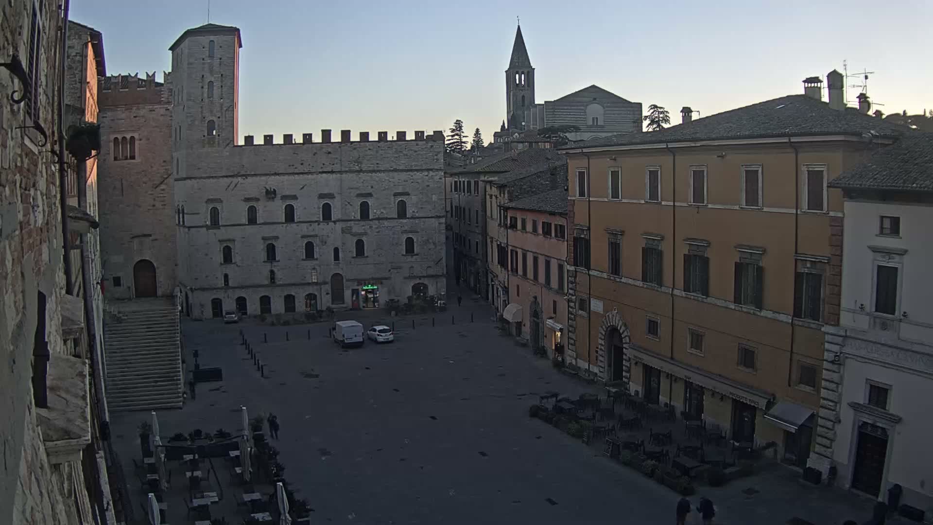 An overcast daytime view reveals a historic European town square with ancient stone buildings, a prominent tower on the left, a distant church spire, and a few people walking across the plaza while an outdoor cafe operates on the right.