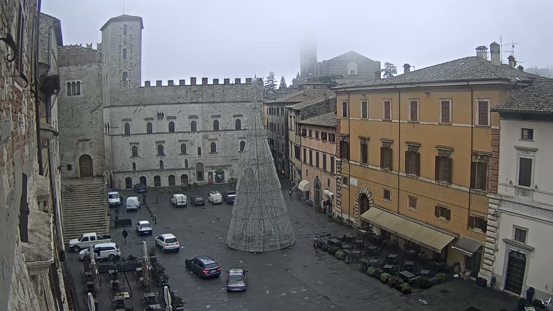 A wide view of a European town square on an overcast and foggy day shows a large, conical decorative structure, surrounded by historic stone buildings and multi-story structures with outdoor cafes, populated by several cars and pedestrians.