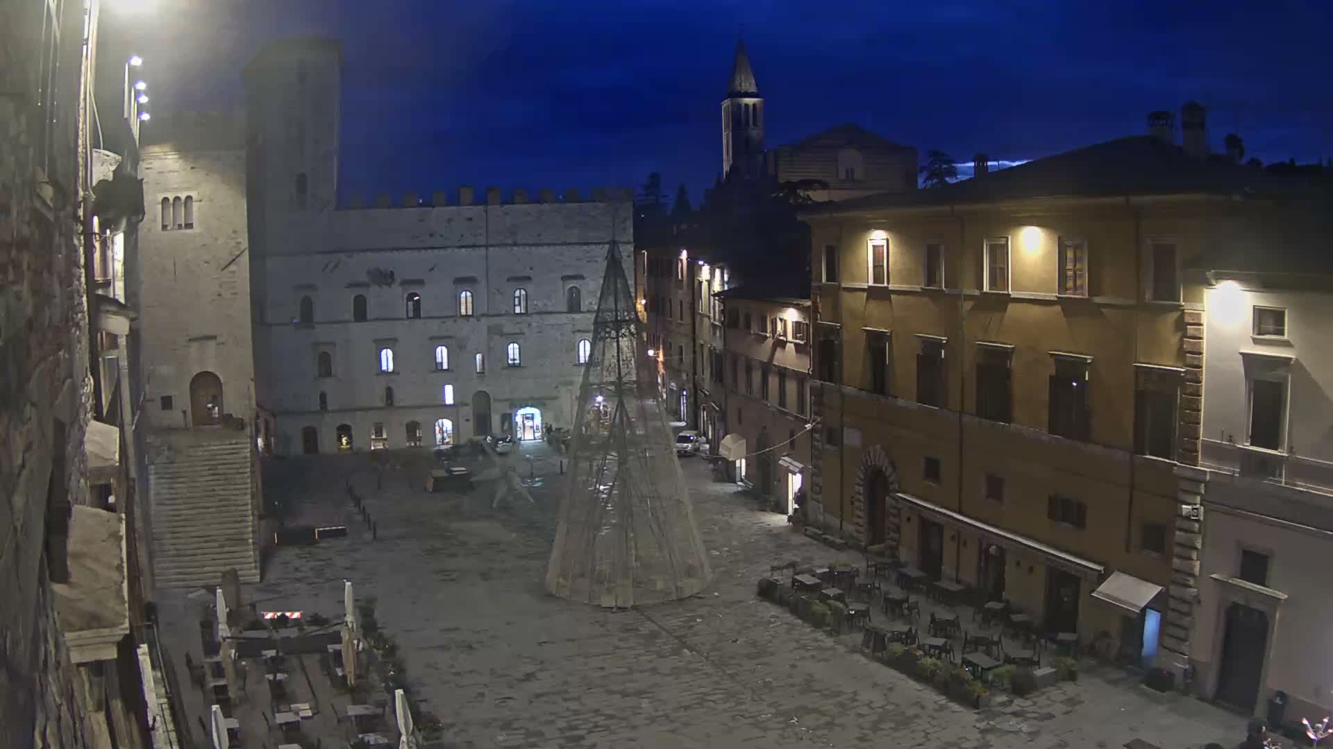 A quiet European town square is seen at night under a clear dark blue sky, centered by a modern conical festive tree, and surrounded by historic buildings with illuminated windows and outdoor cafe seating.