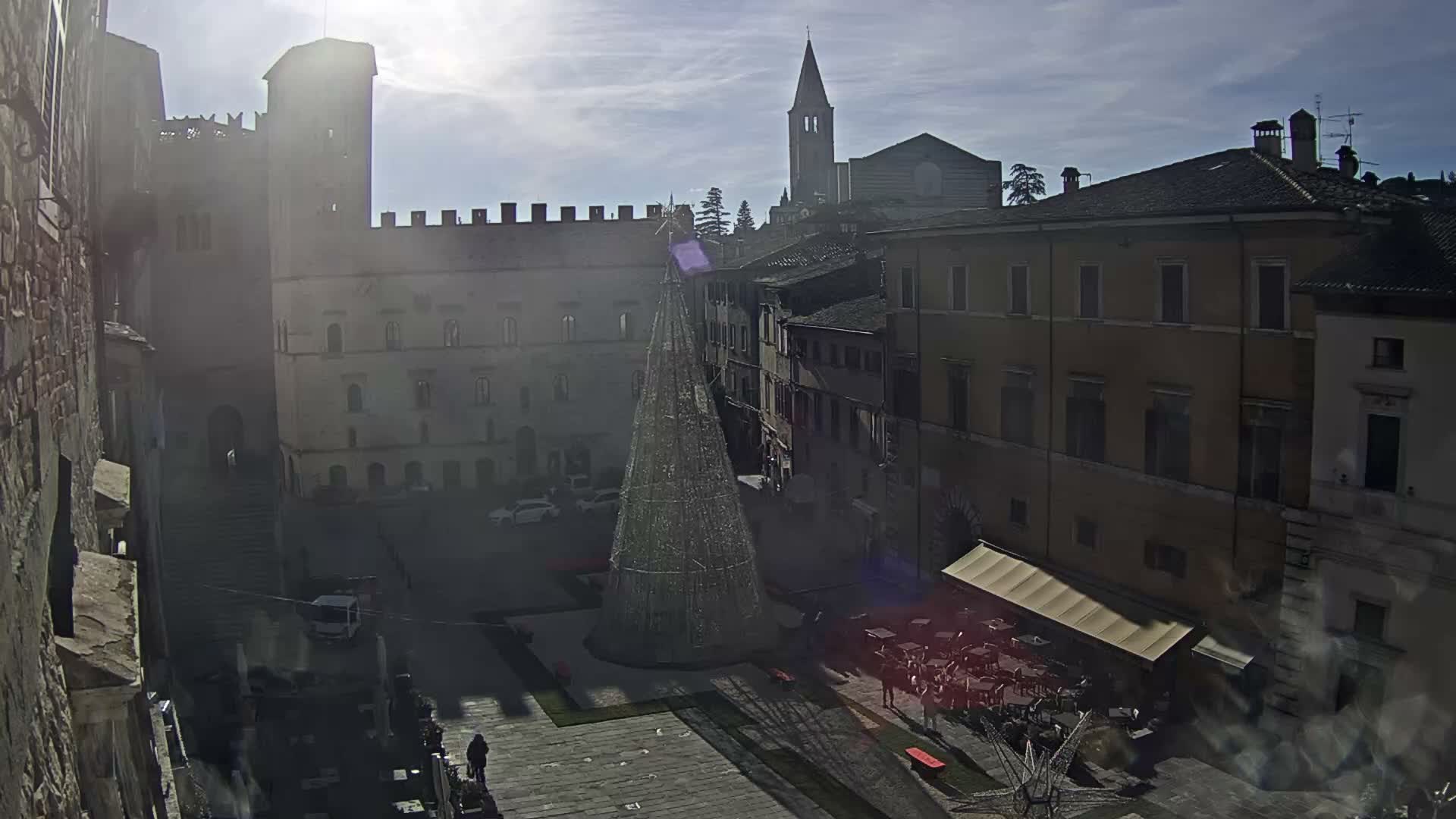 An aerial view reveals a sunlit European town square featuring a large illuminated Christmas tree, historic buildings, an active outdoor cafe, and a prominent church tower under a partly cloudy sky.