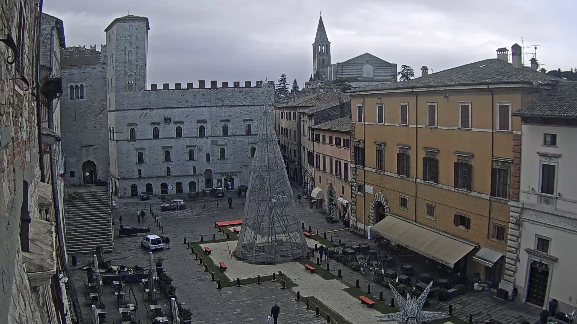 An aerial view reveals a sunlit European town square featuring a large illuminated Christmas tree, historic buildings, an active outdoor cafe, and a prominent church tower under a partly cloudy sky.