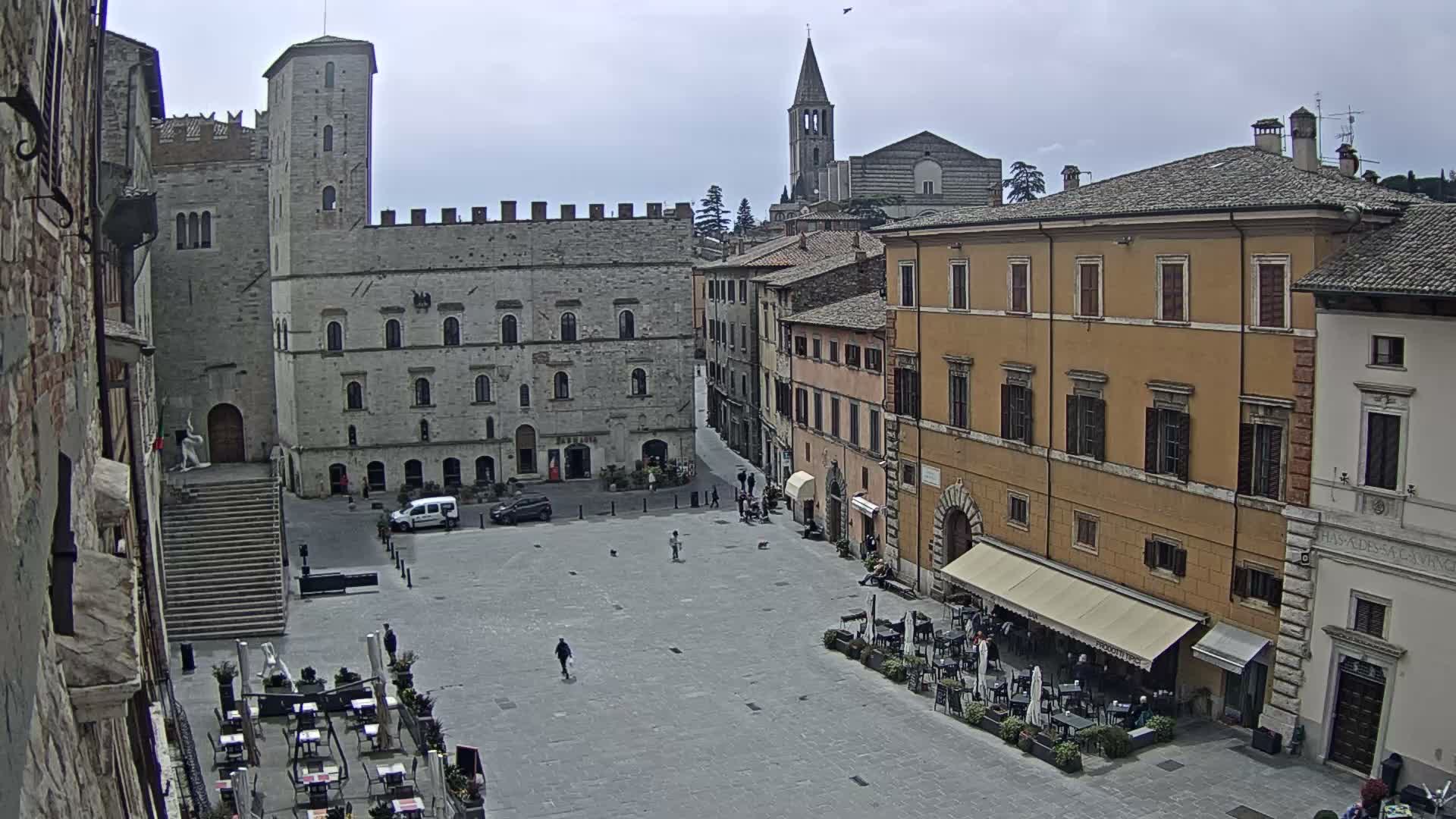 An aerial view reveals a sunlit European town square featuring a large illuminated Christmas tree, historic buildings, an active outdoor cafe, and a prominent church tower under a partly cloudy sky.