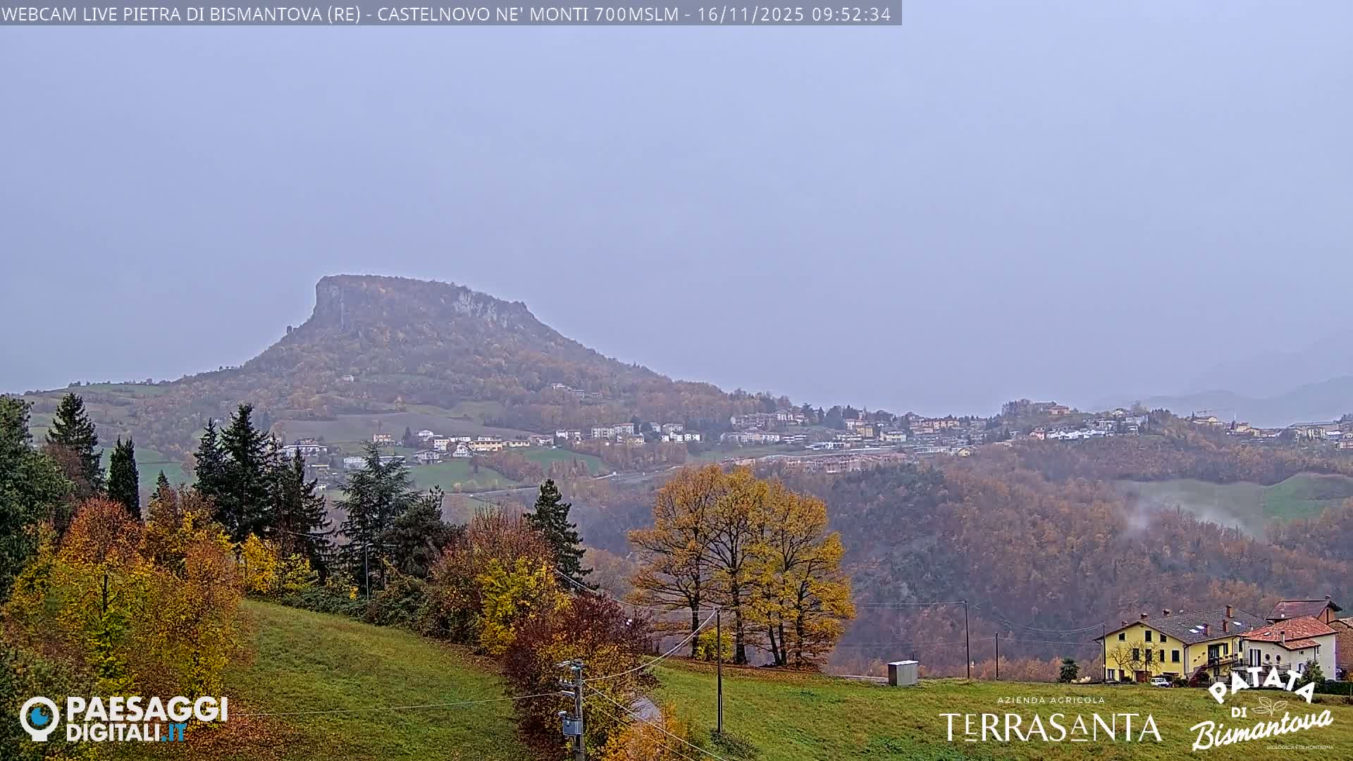 An overcast day reveals a broad landscape with a prominent flat-topped mountain, a village nestled among rolling hills displaying rich autumn foliage, and scattered houses in the foreground.