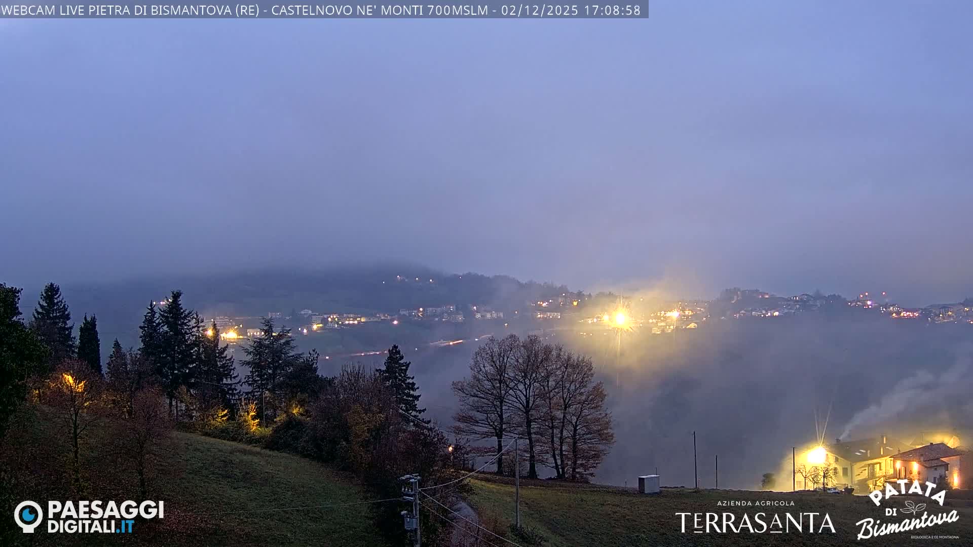 A misty or foggy twilight view shows a village nestled in a valley among hills, with scattered lights illuminating buildings and roads through the haze.